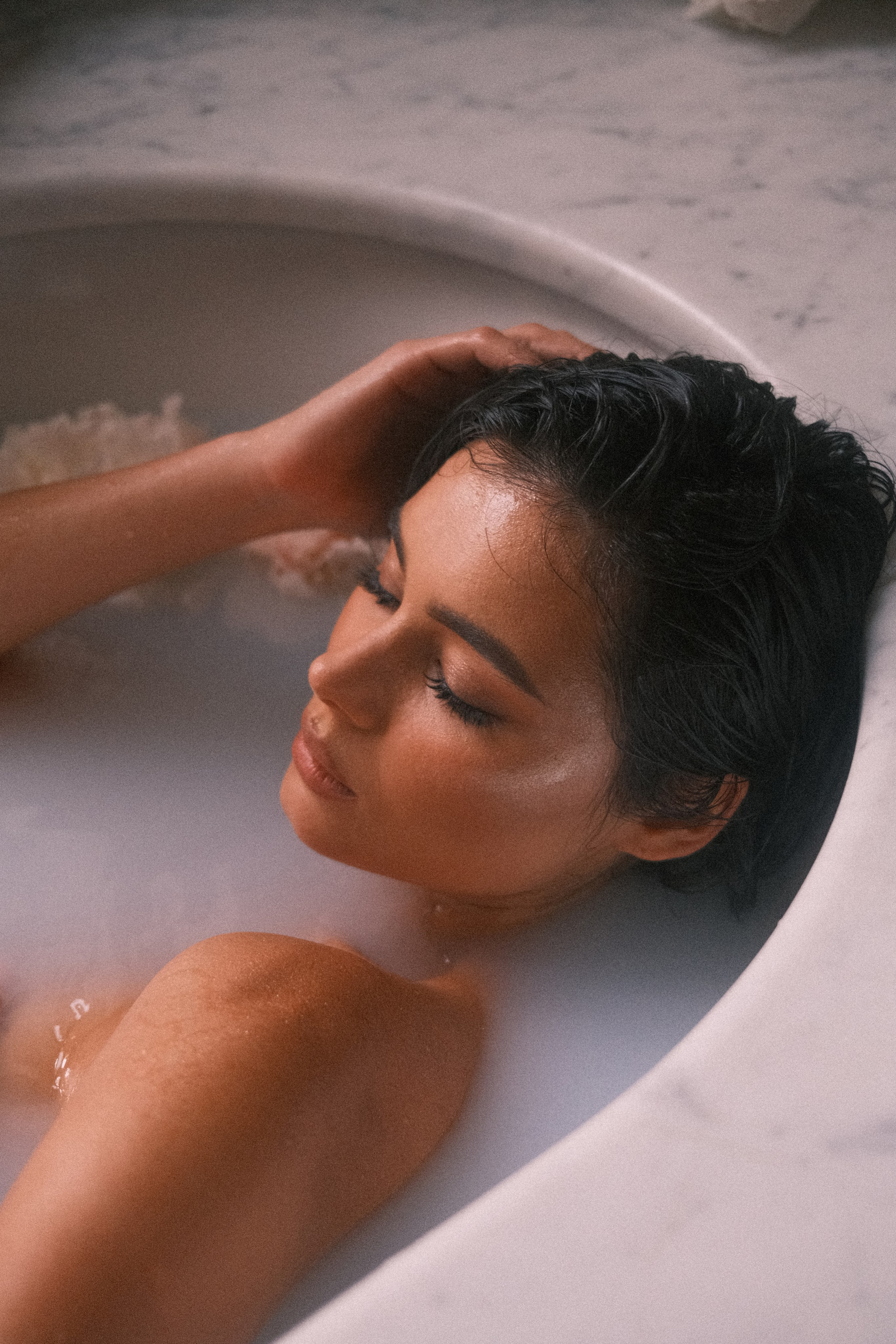 A tranquil close-up of a woman reclining in a marble bathtub, her hand resting on her head and her face exuding calmness.