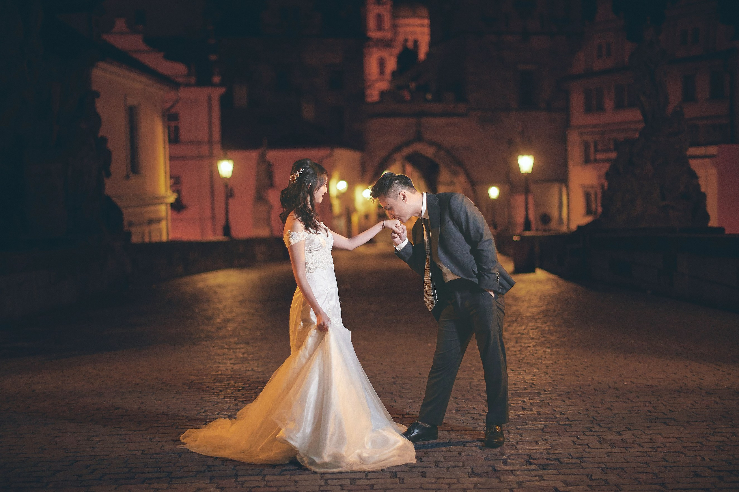 Groom Conan kissing bride Eva’s hand tenderly on Charles Bridge at dawn, Prague.