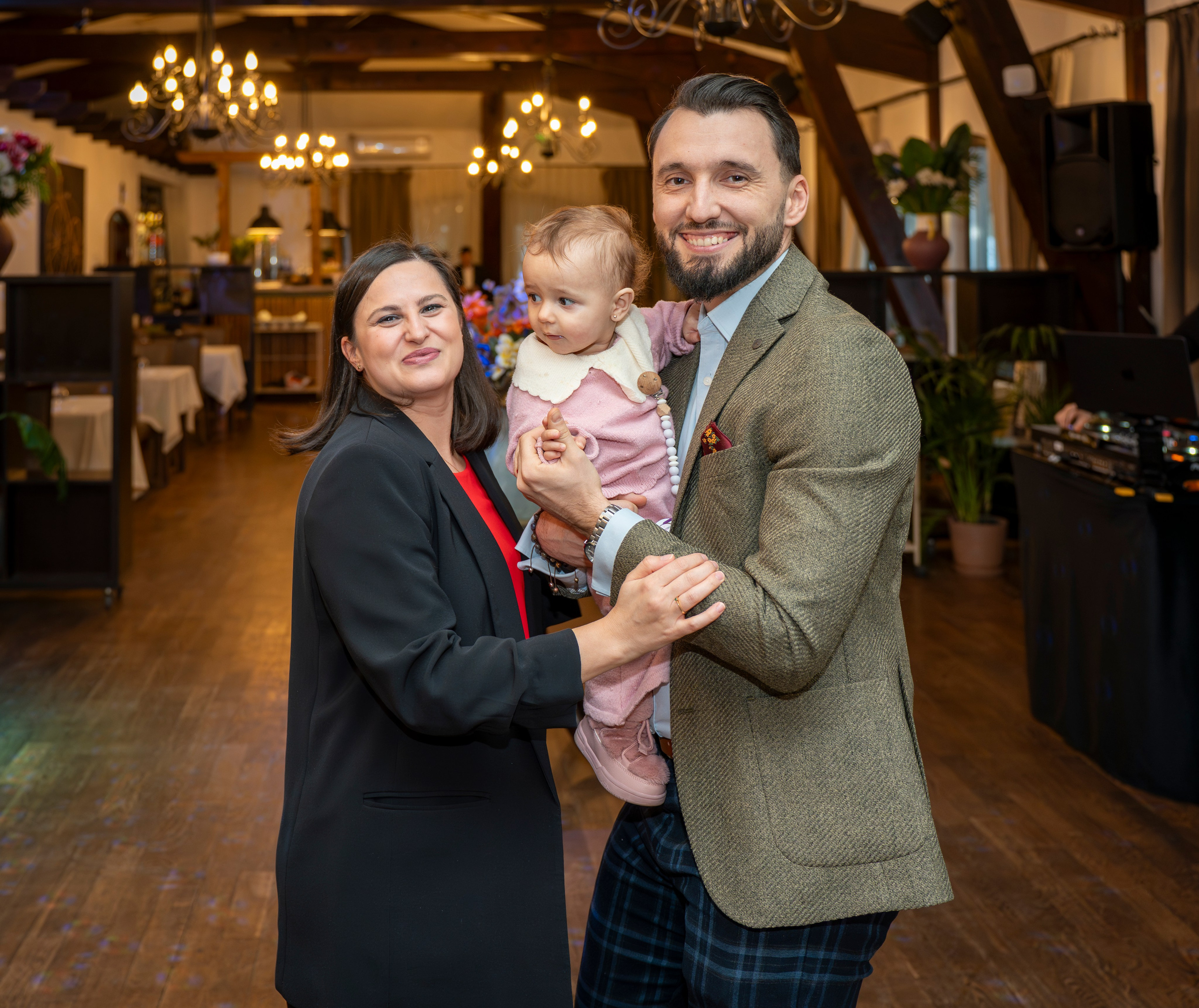 Man and woman holding a baby in an elegant indoor venue with chandeliers and wooden ceiling beams.