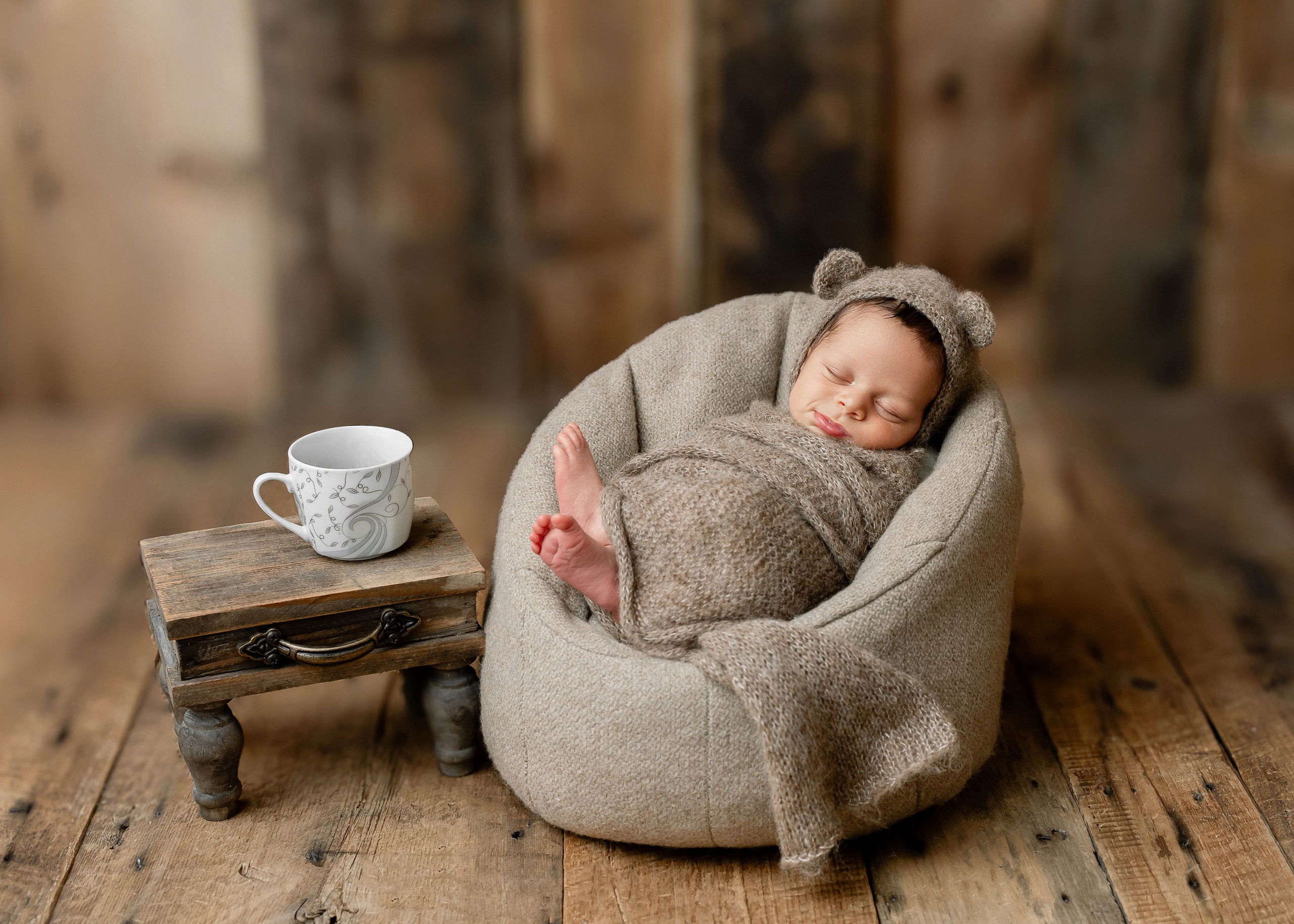 Newborn baby wrapped in a cozy brown swaddle, sleeping in a bear-ear outfit on a soft beanbag chair with a rustic wood background and a tiny side table with a teacup — whimsical and warm newborn photography setup.