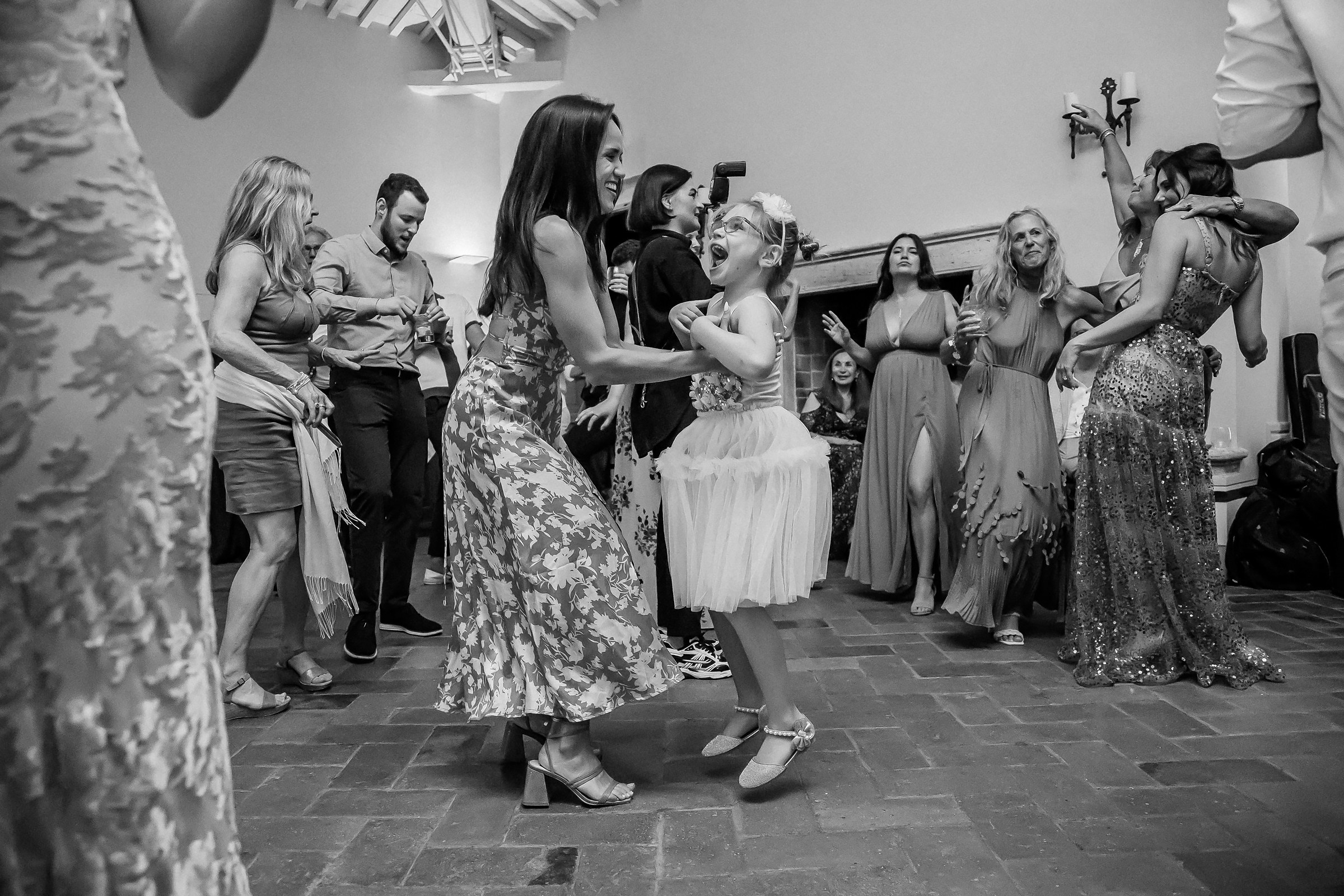 A woman in a floral dress dancing joyfully with a young girl in a white tutu, as other guests enjoy the music in the background.