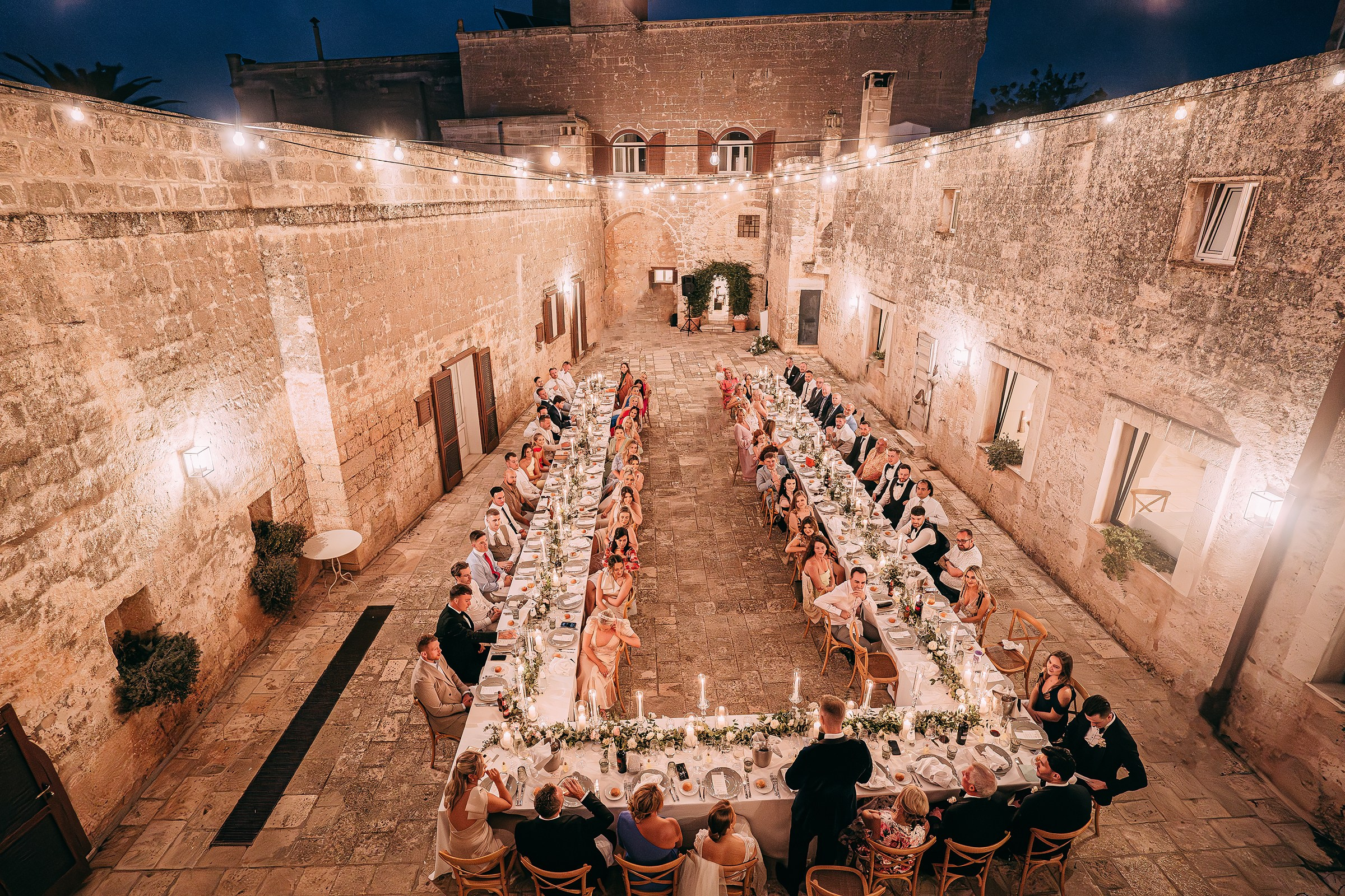 A wide-angle black-and-white shot of the groom delivering a heartfelt speech, with the entire wedding party attentively seated around the banquet table.
