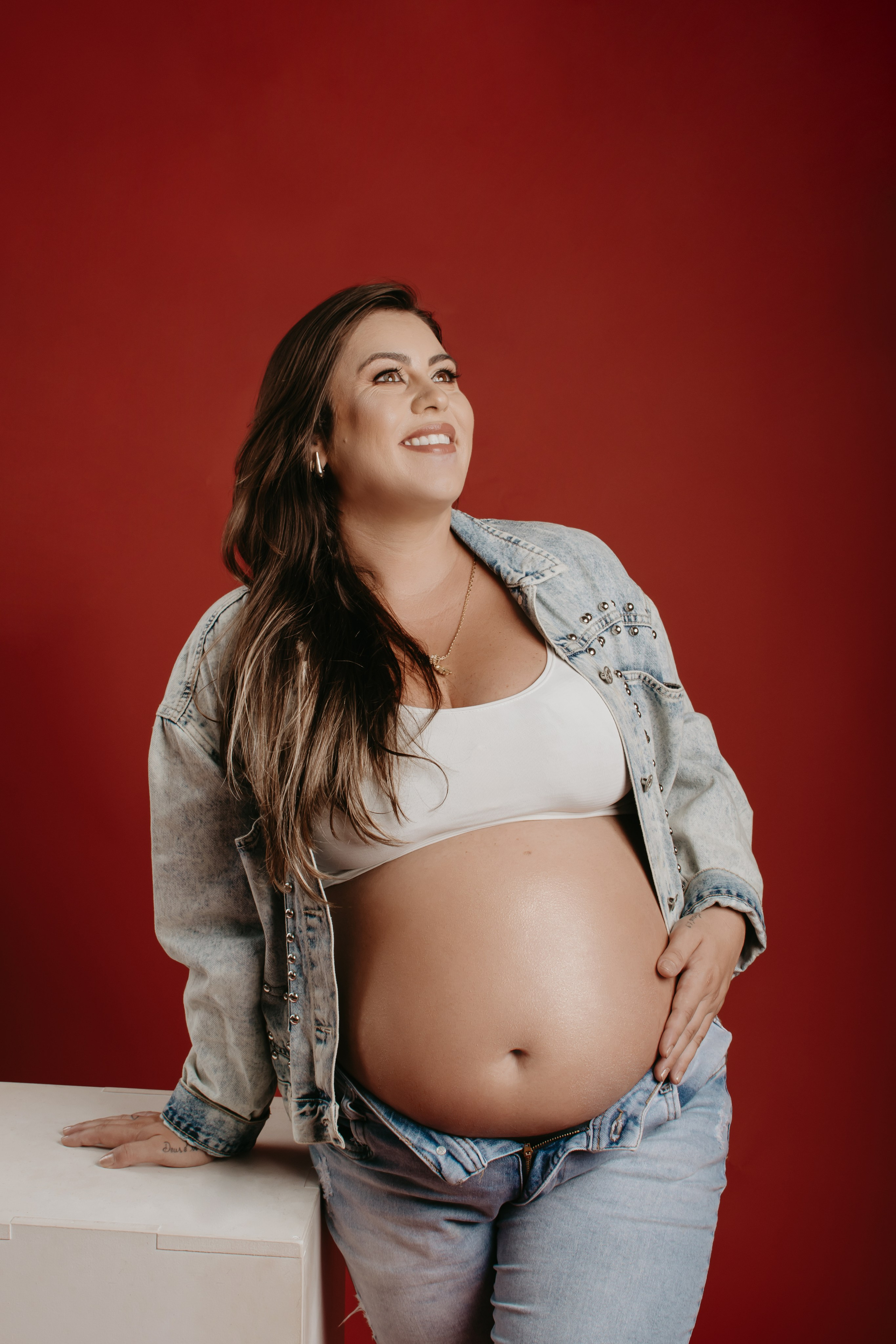 Foto de uma gestante empoderada em fundo colorido vermelho com roupas jeans, que remetem a um look moderno e atemporal