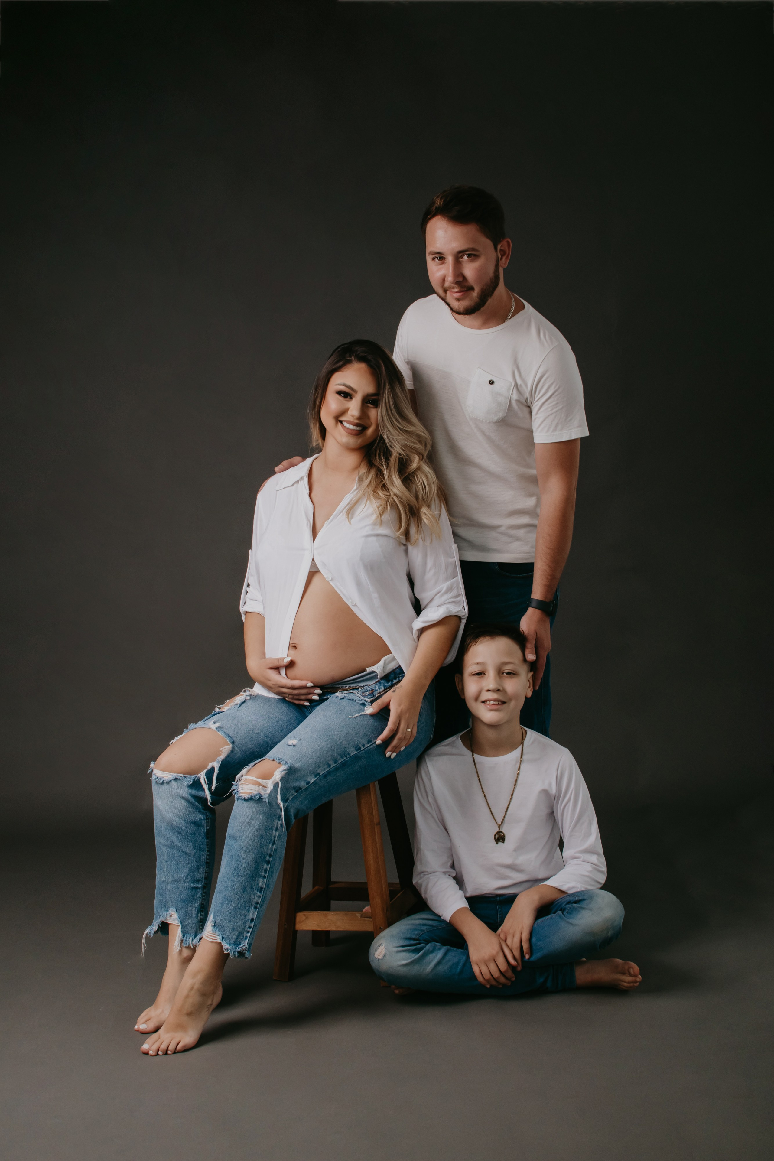 Ensaio gestante em estúdio com família, vestindo jeans e camisa branca, expressando união e alegria à espera do bebê