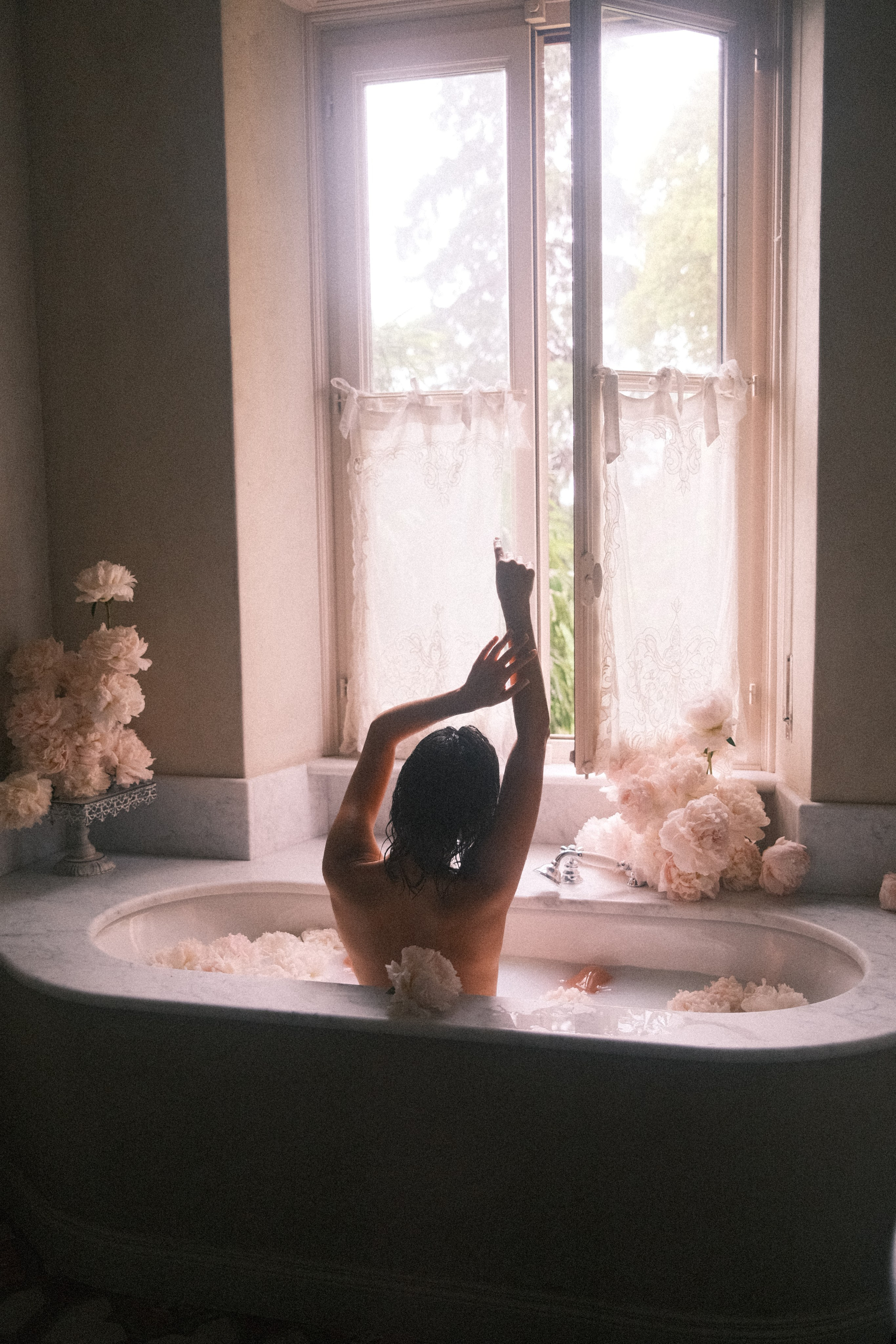 A backlit image of a woman stretching her arms in a marble bathtub, framed by a large window with soft light streaming through lace curtains and surrounded by floral decor.