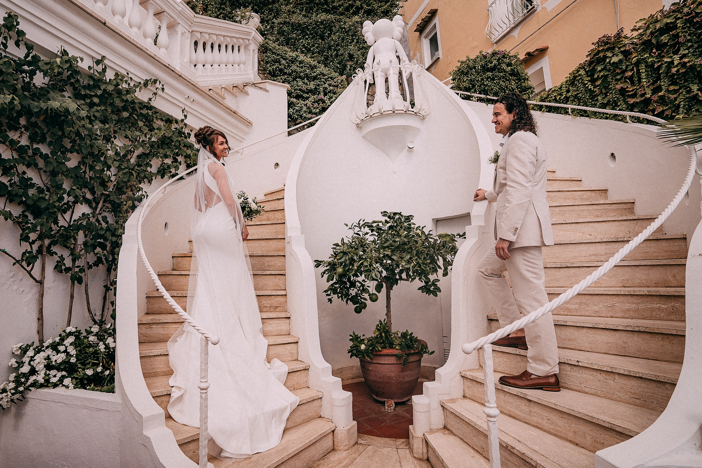 A bride and groom on a white spiral staircase, surrounded by lush greenery and Mediterranean architecture.