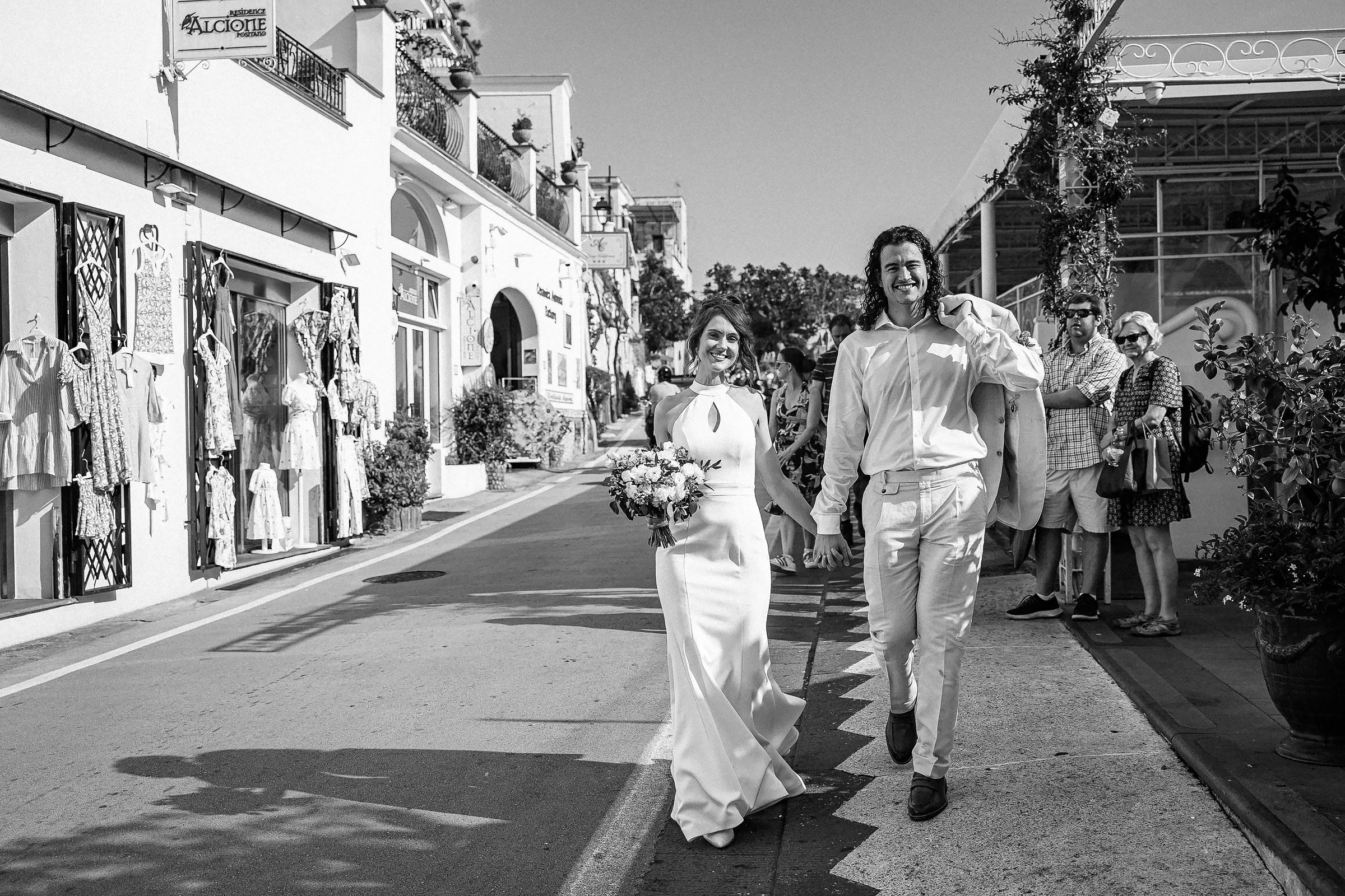 A black-and-white photo of a newlywed couple walking through the streets of Positano, surrounded by locals and tourists, with boutique shops lining the road.