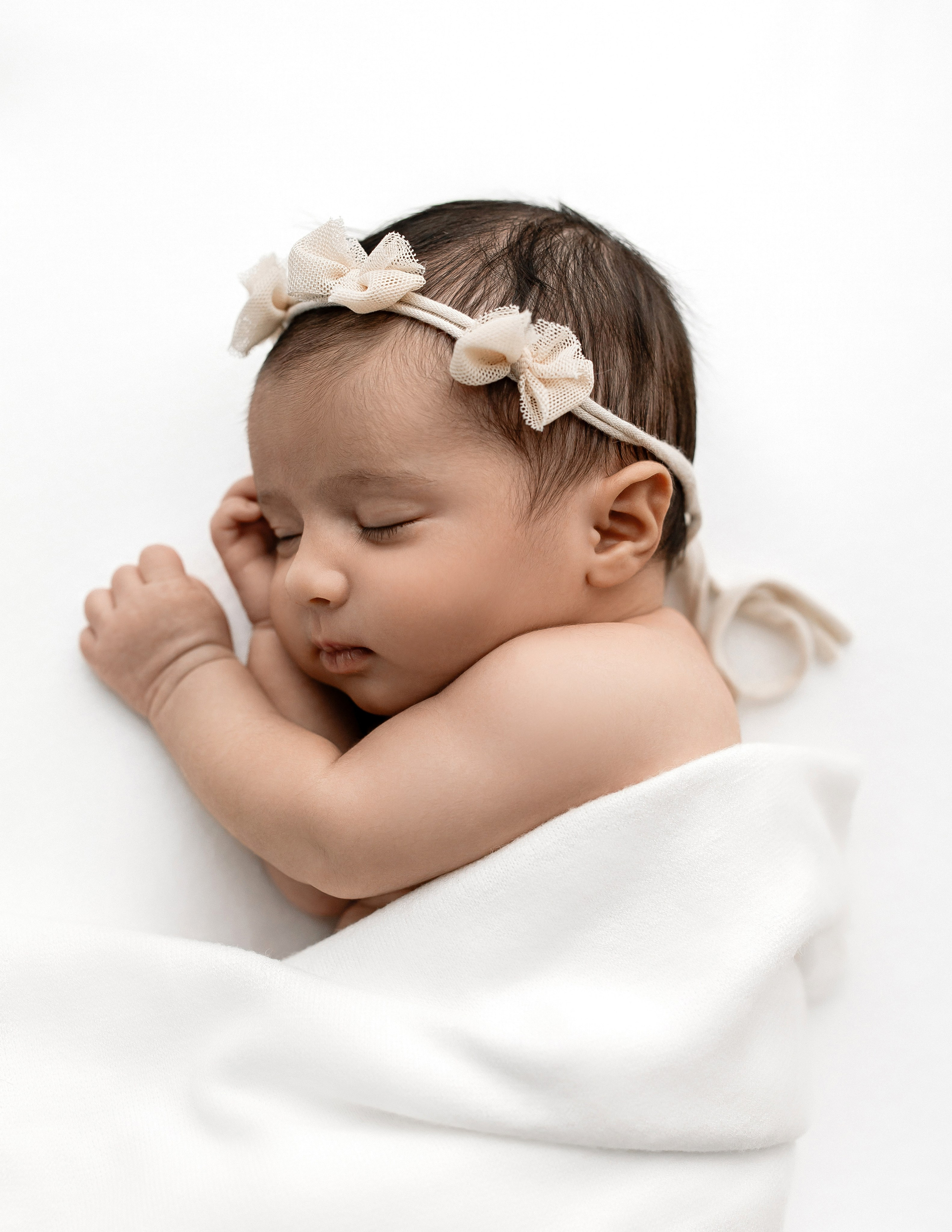 Newborn baby girl sleeping peacefully on a white blanket, wearing a soft beige floral headband — professional minimalist newborn photography.