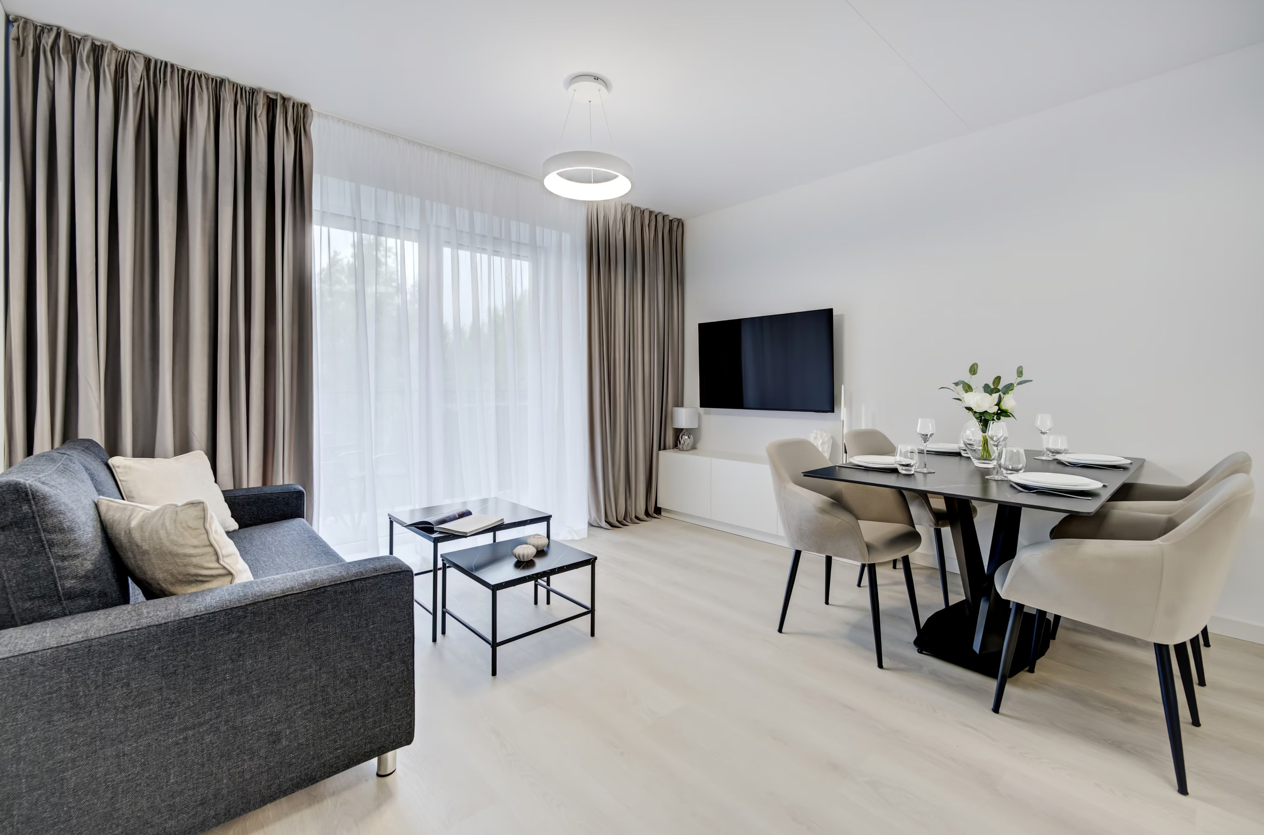 Stilinga svetainė su tamsia sofa, juodais staliukais ir elegantiška valgomojo zona.