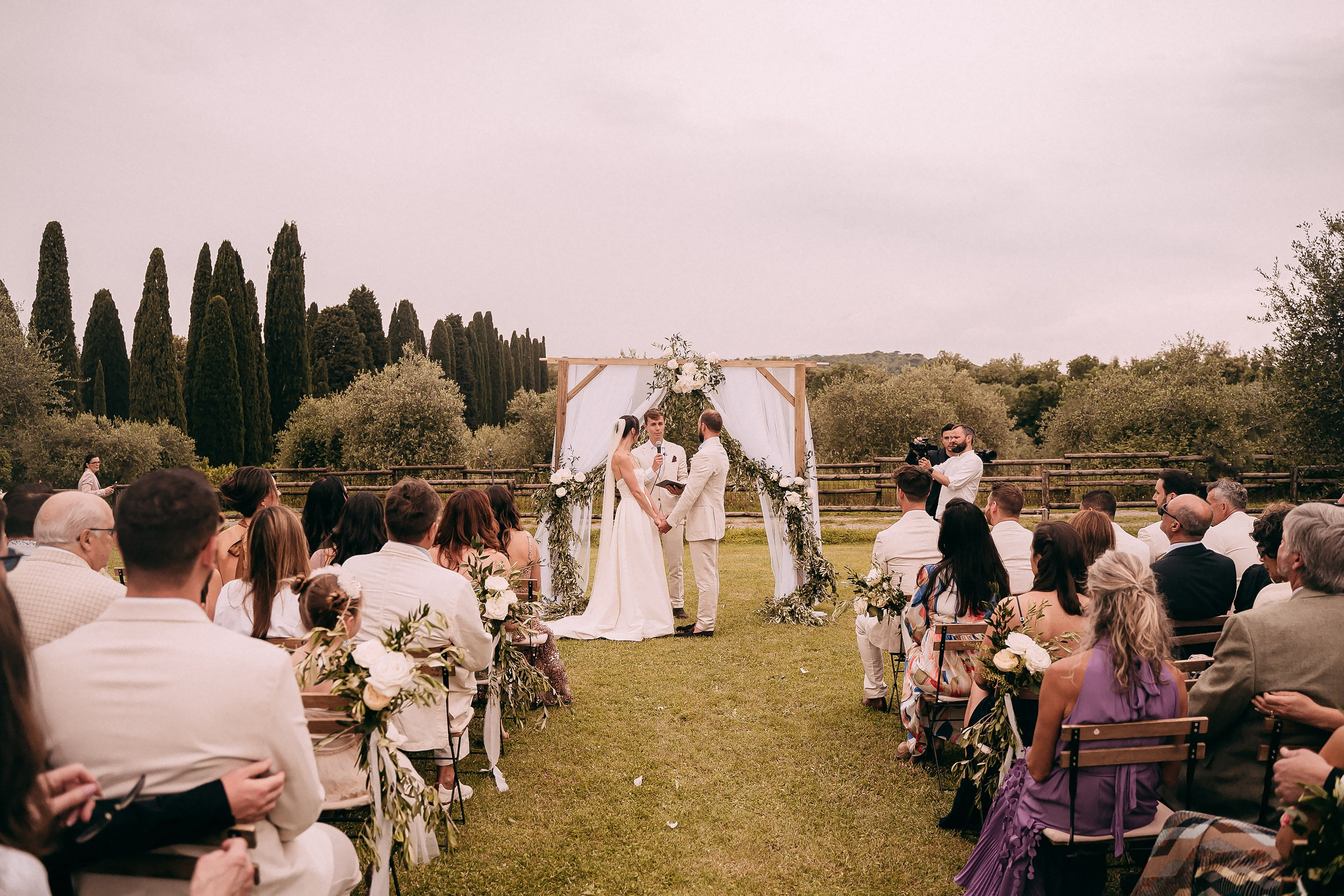 The couple stands under a decorated wooden arch, holding hands, exchanging vows amidst a serene outdoor setting.