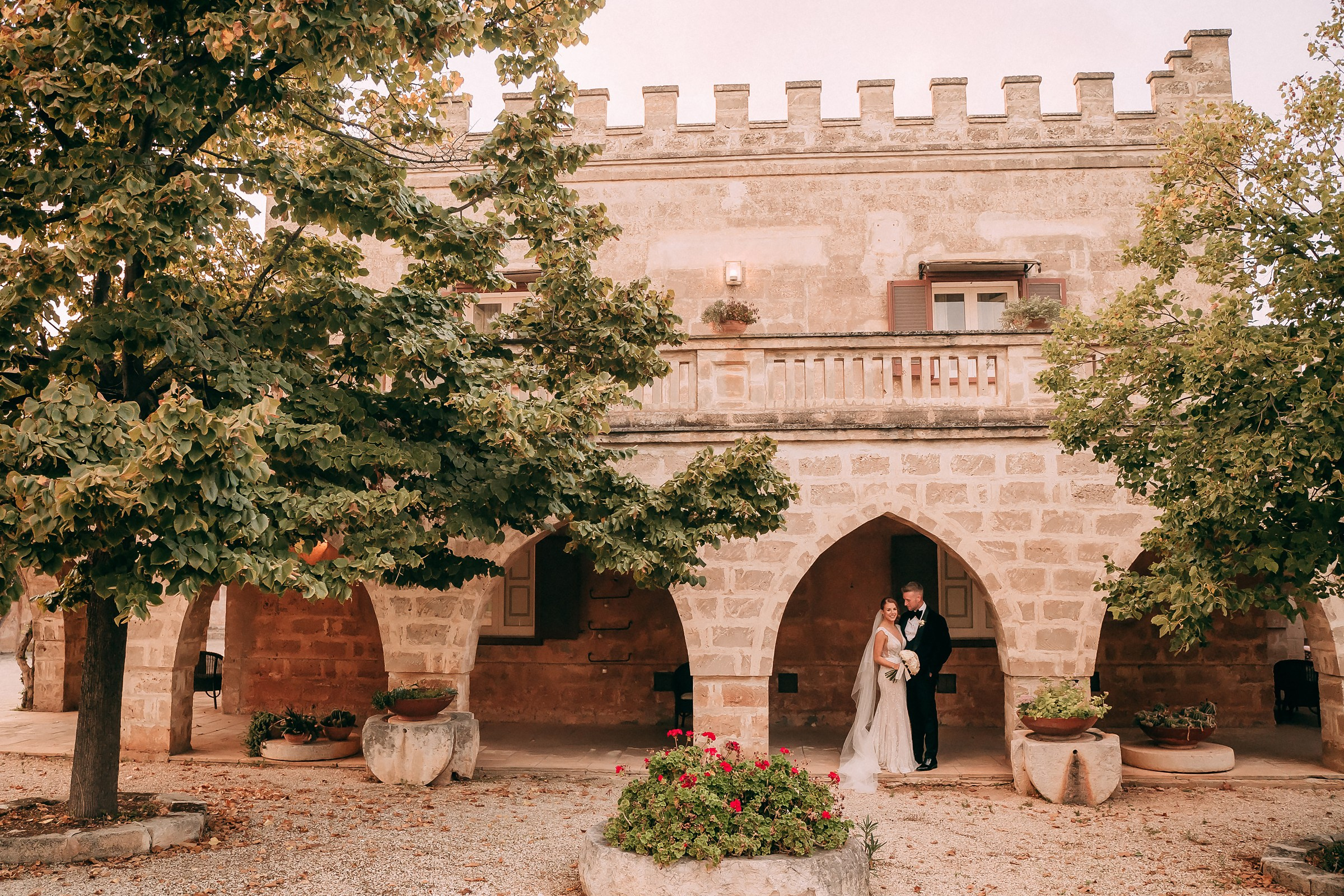 A newlywed couple poses under a historic stone building’s archway, the bride holding her bouquet, blending modern romance with timeless architecture.