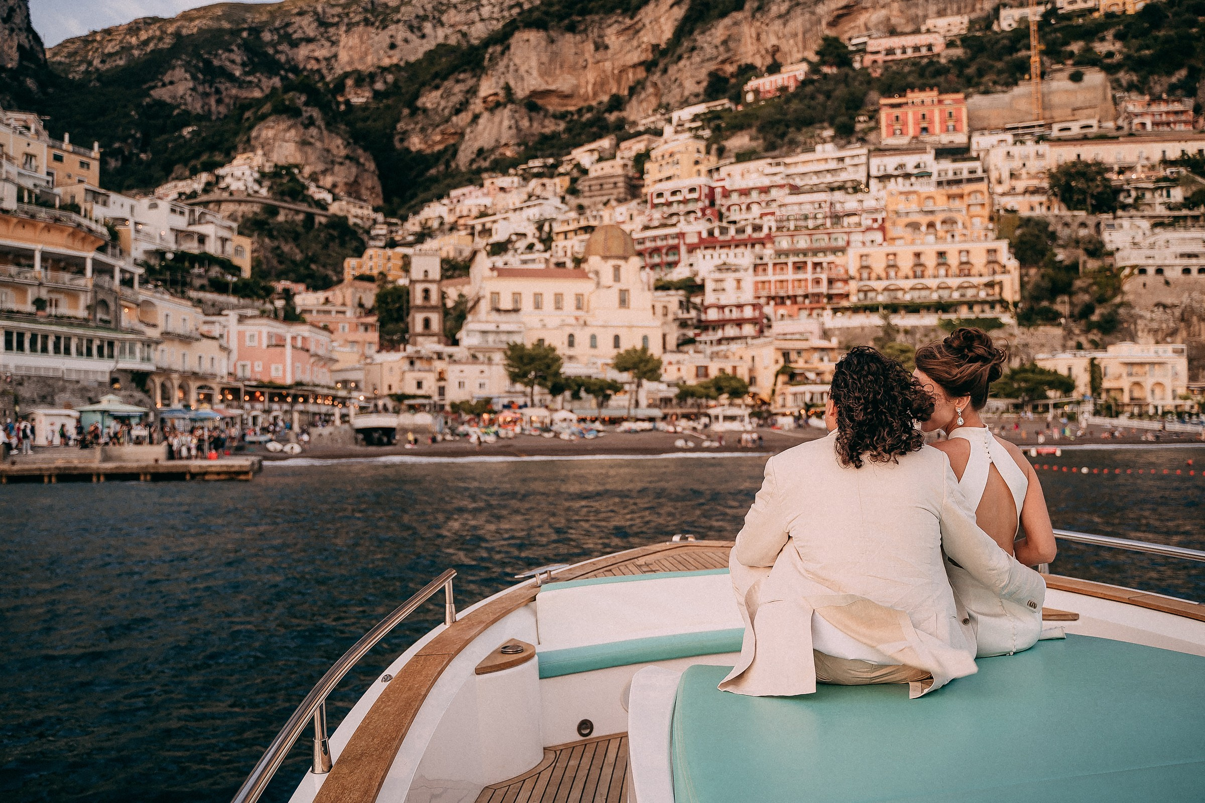 The couple gazes at the picturesque Positano coastline from the boat, with its colorful houses cascading down the cliffside toward the sea.
