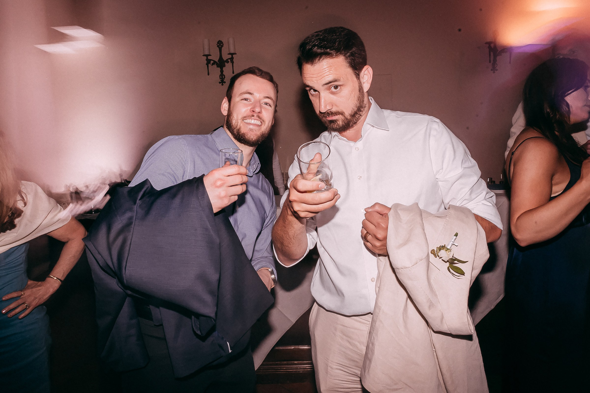 Two men posing for the camera at a wedding reception, each holding a glass, one pointing playfully, while dressed in semi-formal attire.