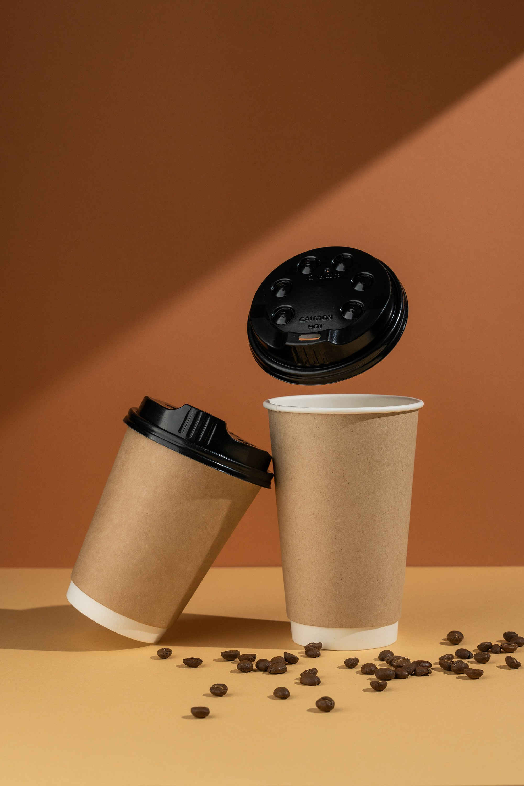 Açıq qəhvəyi fonda qara qapaqlı iki ədəd qəhvəyi kağız kofe stəkanı. Stəkanlardan biri əyri durur, digəri isə dikdir və qapağı stəkanın üstündə havada süzülür. Stəkanların ətrafına bir neçə qəhvə dənəsi səpələnib. Arxa fonda işıq kölgə salır.