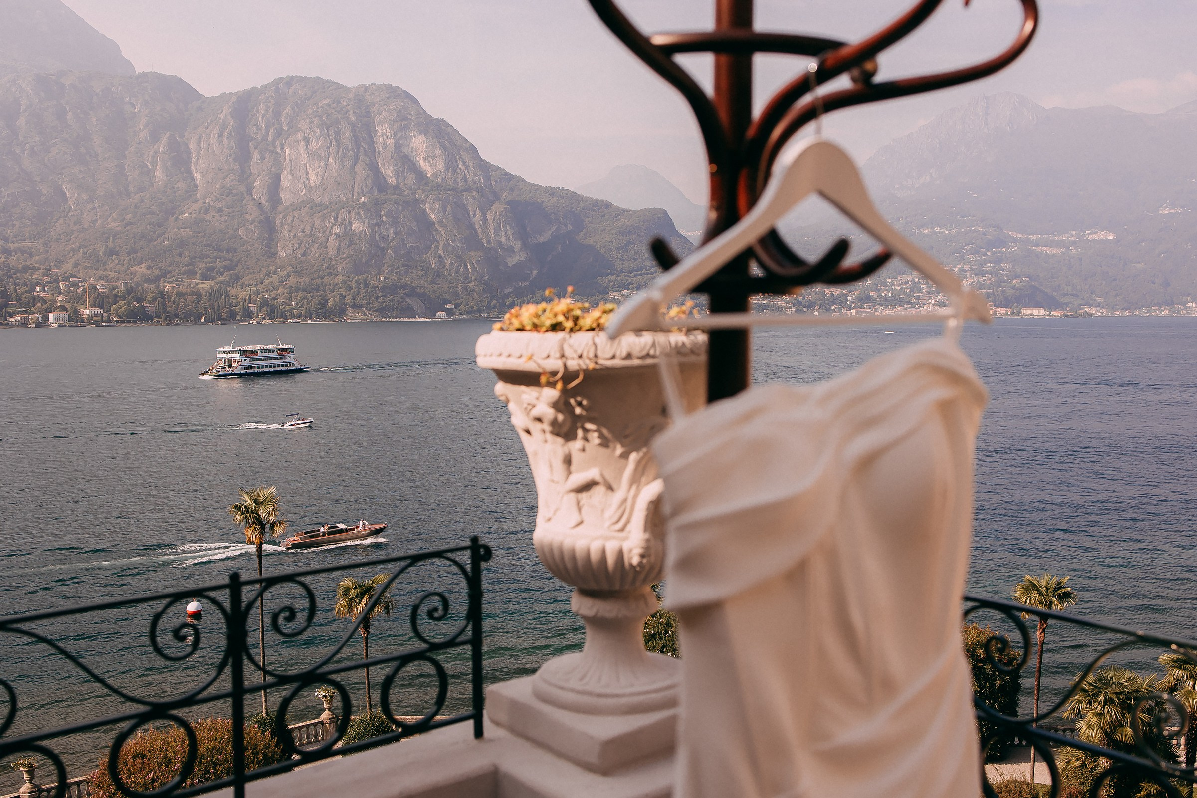 A wedding dress hanging on a coat stand on a terrace, framed by the scenic view of Lake Como’s sparkling waters and majestic mountains.