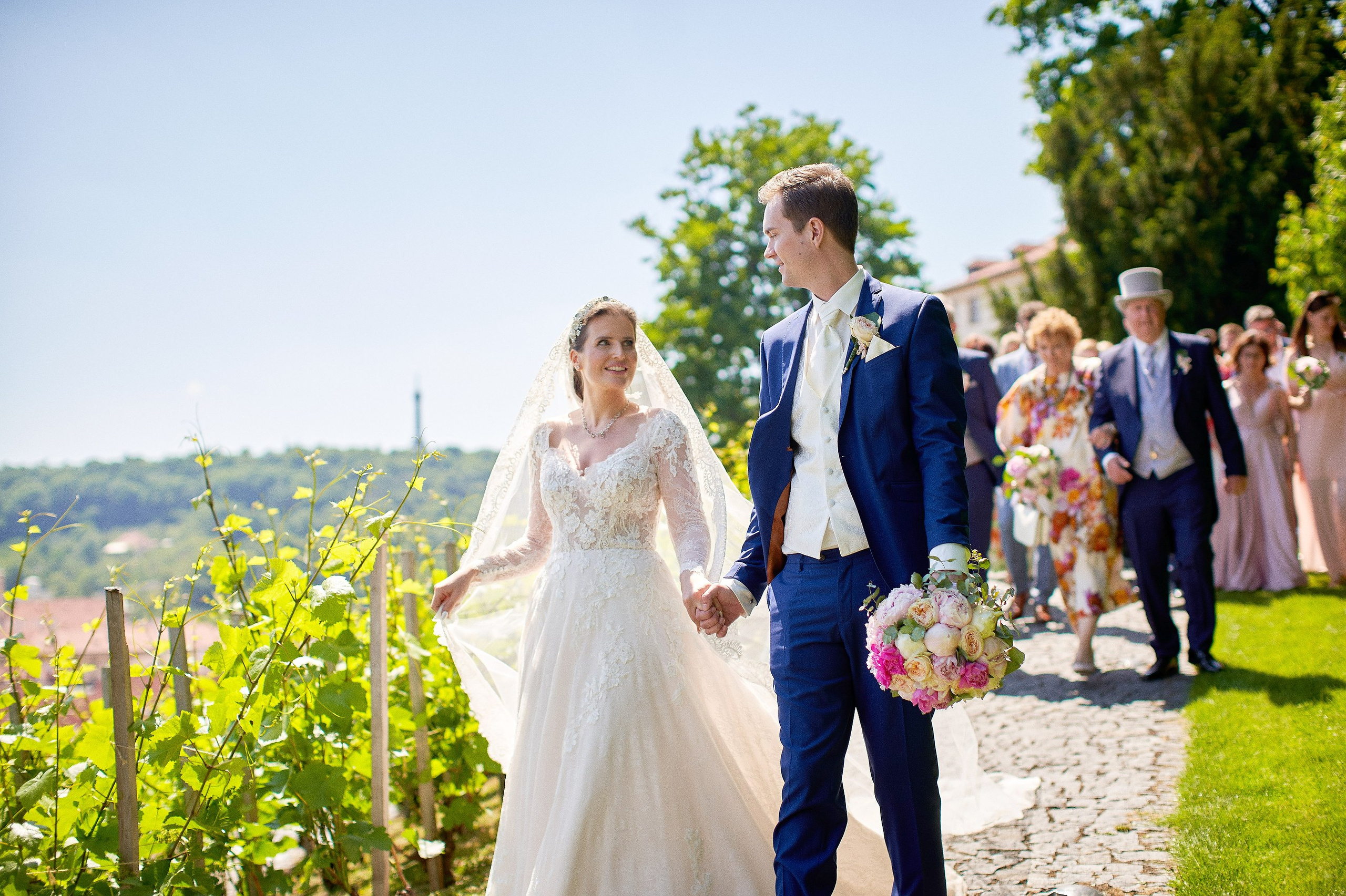 A smiling, happy bride & groom hold hands as they walk through the vineyards overlooking the city of Prague down below.