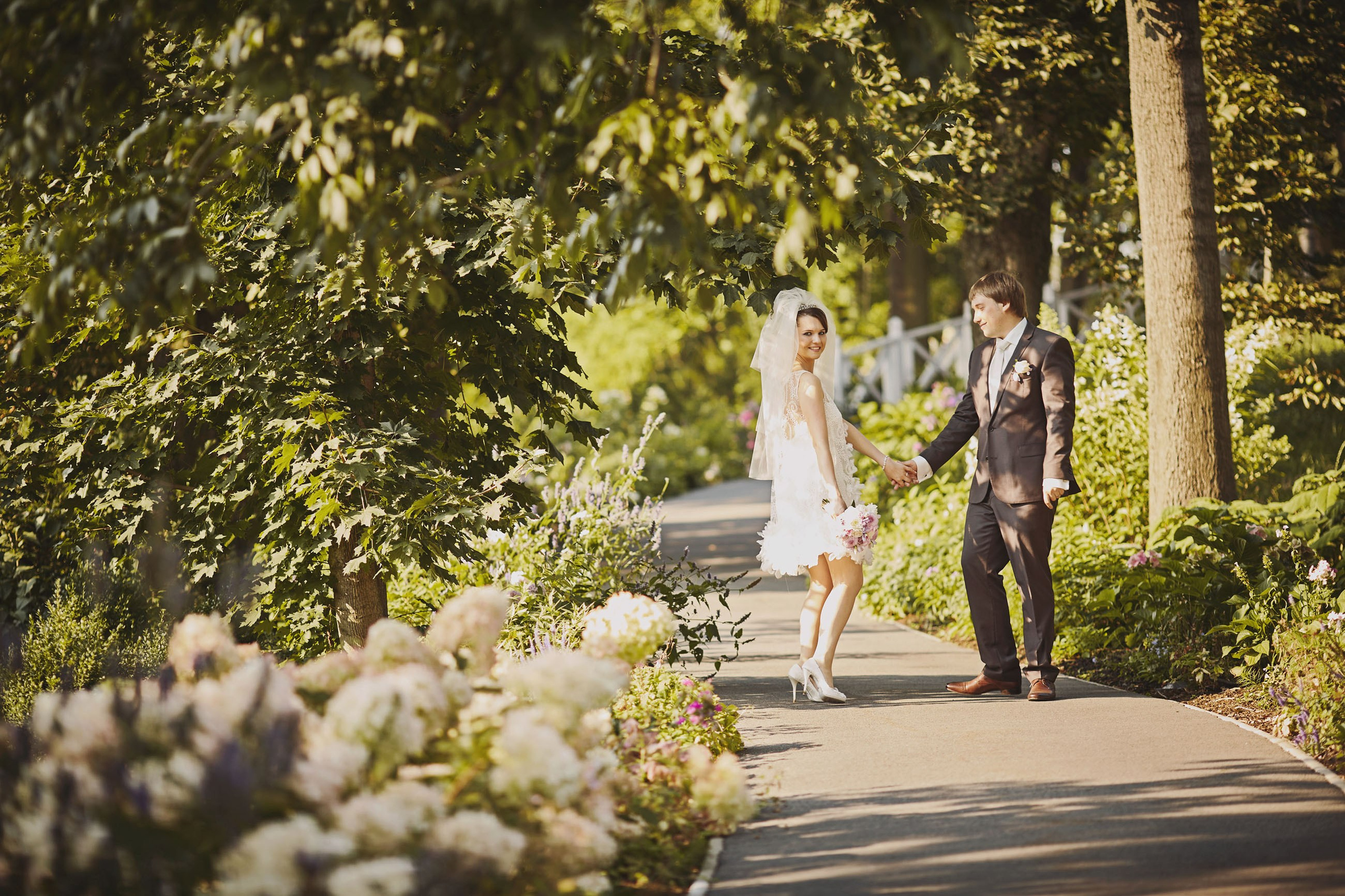 Newlyweds watlk under the trees on a flower-lined path on the grounds of the Chateau Mcely following their outdoor wedding ceremony.