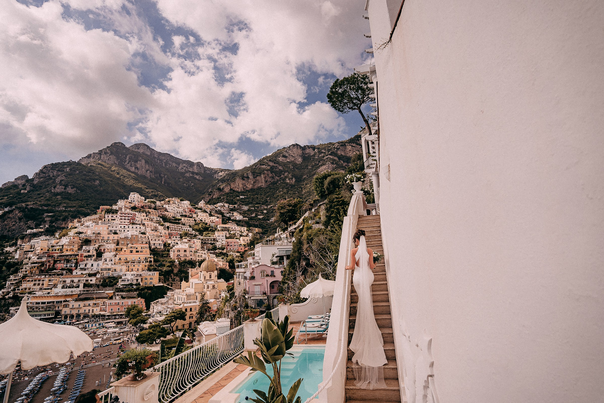A bride walking up a staircase beside a pool, her long wedding dress flowing behind her, with the beautiful skyline in the background.