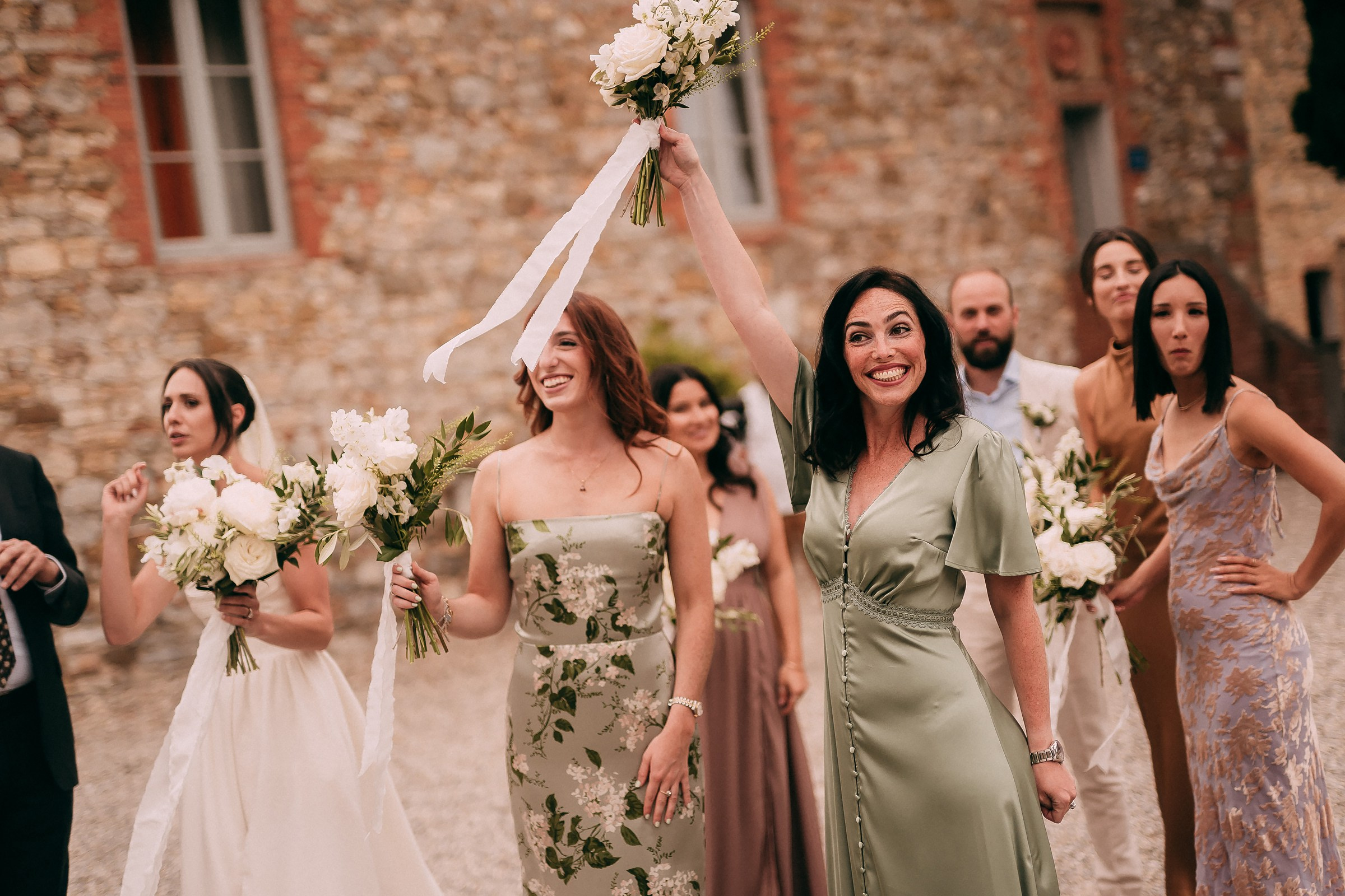 Bridesmaid triumphantly raising bouquet, surrounded by a joyful bridal party outside a rustic Tuscan building.