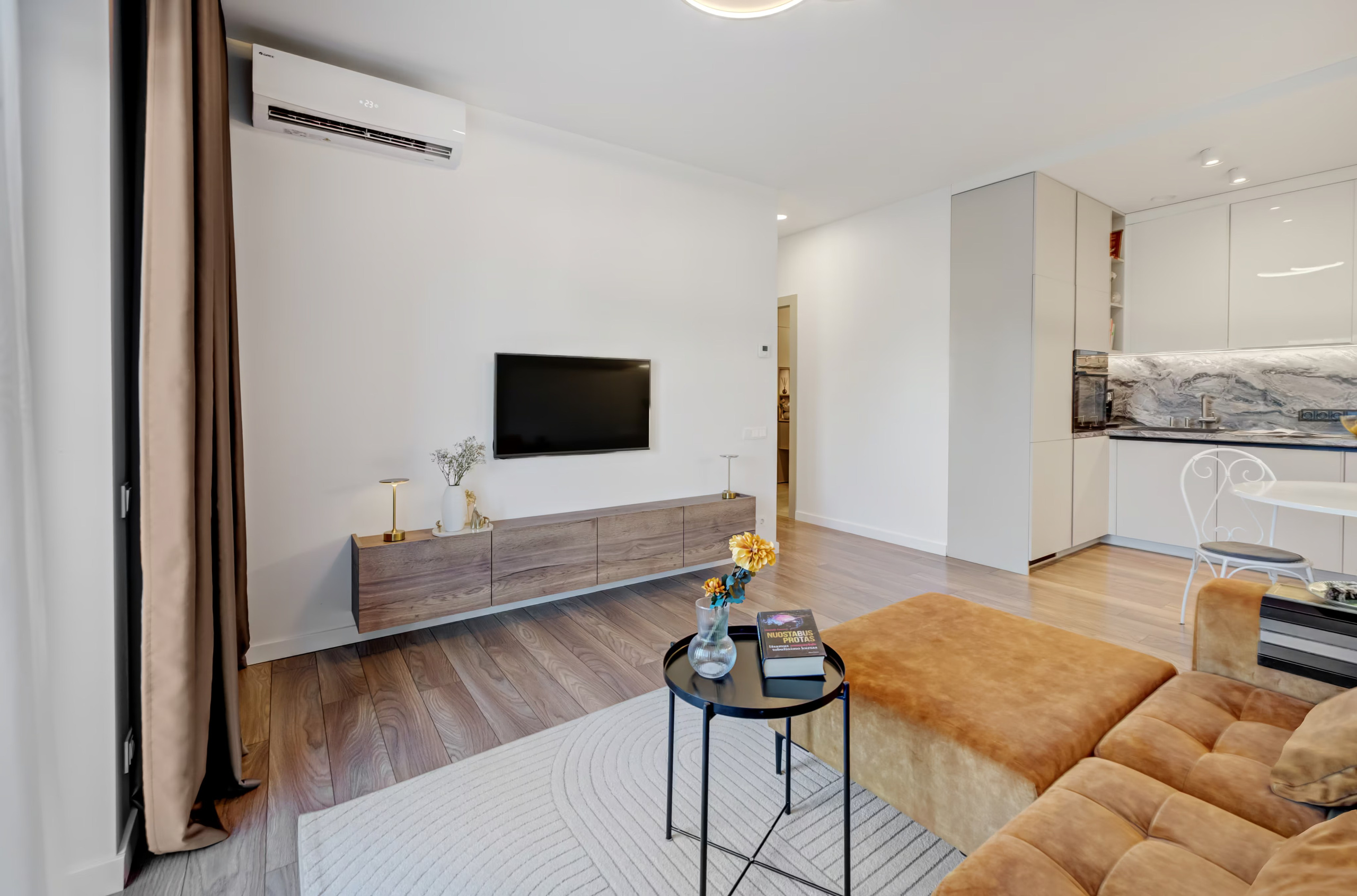 Bendras svetainės vaizdas: moderni sofa, balkono durys su užuolaidomis ir televizoriaus zona.