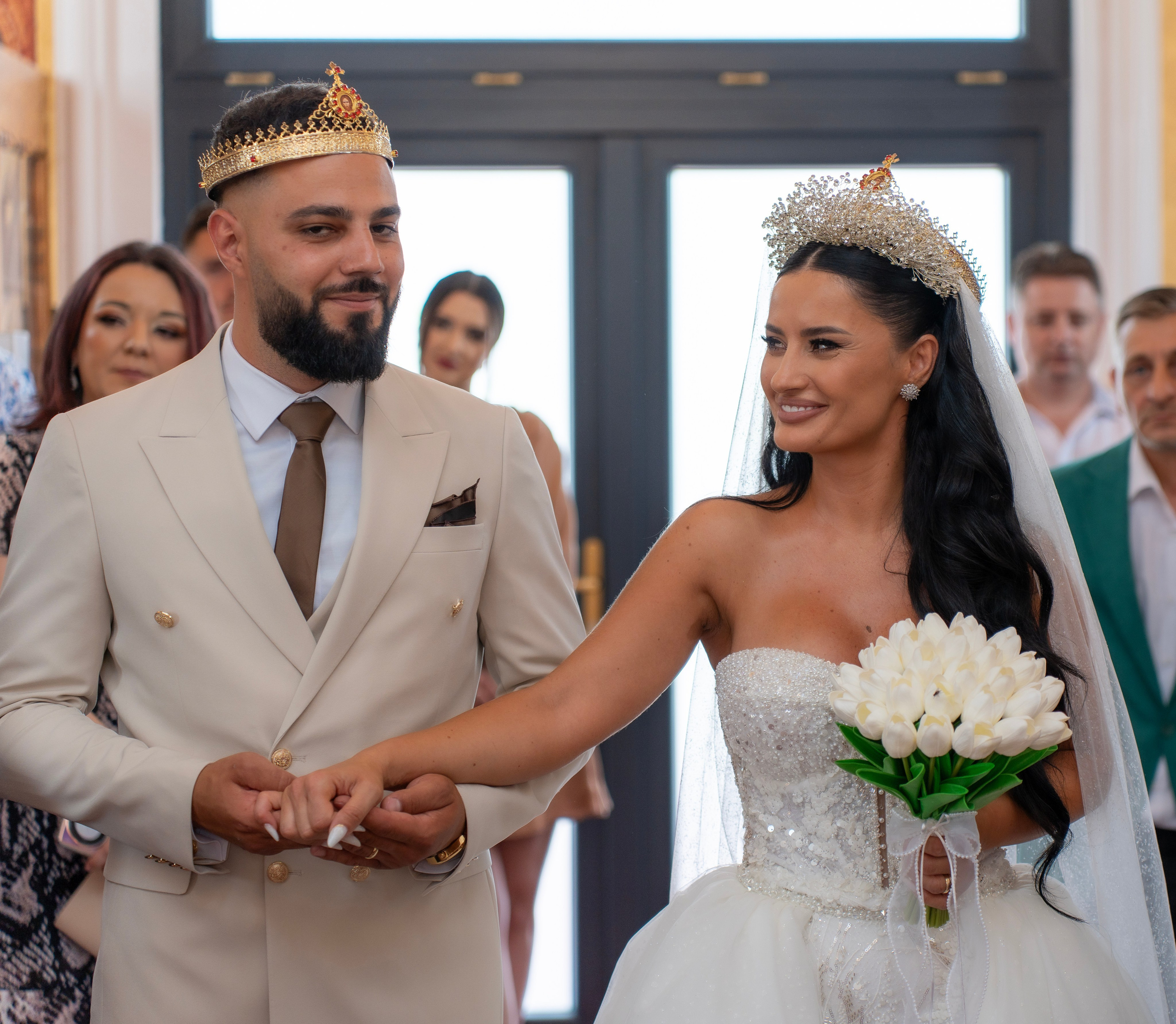 Bride and groom holding hands during wedding ceremony, both wearing crowns. Bride holds a bouquet of white flowers.