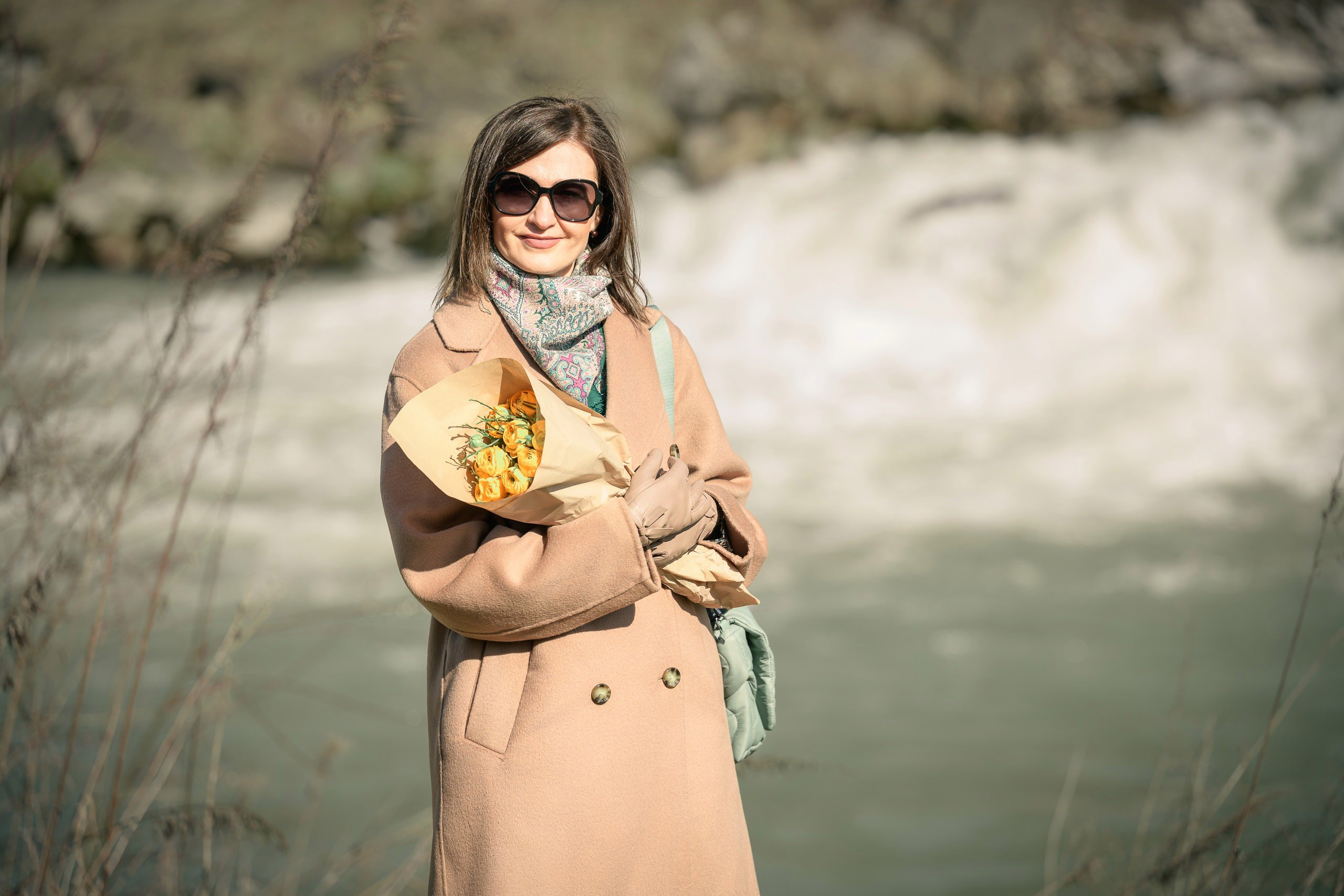 Porträt einer jungen Frau in beigefarbenem Mantel, Handschuhen und Schal vor einem Wald und einem zurückweichenden Fluss.