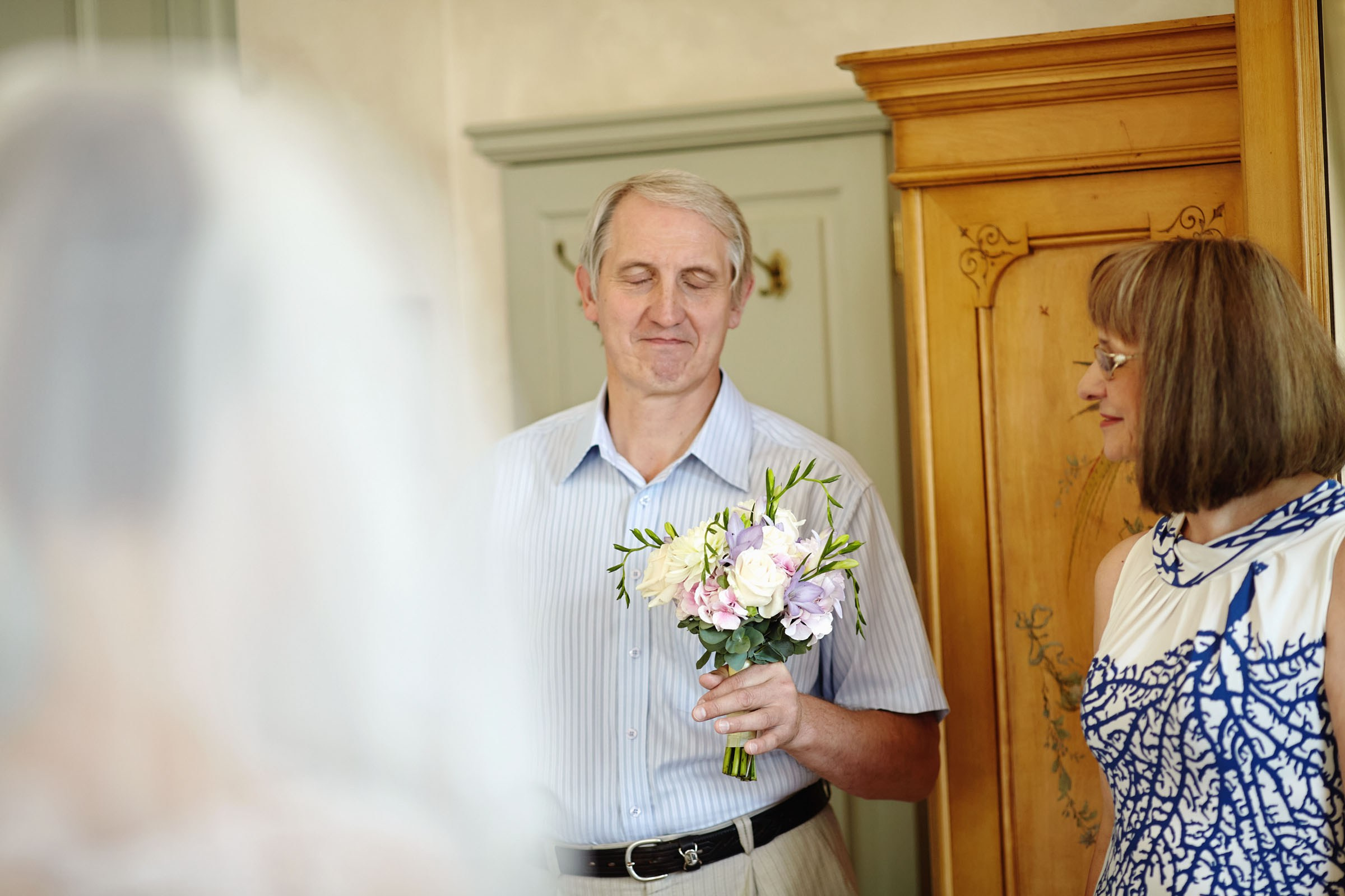 Father of the bride with eyes closed, holding bouquet, as daughter prepares in Chateau Mcely suite.
