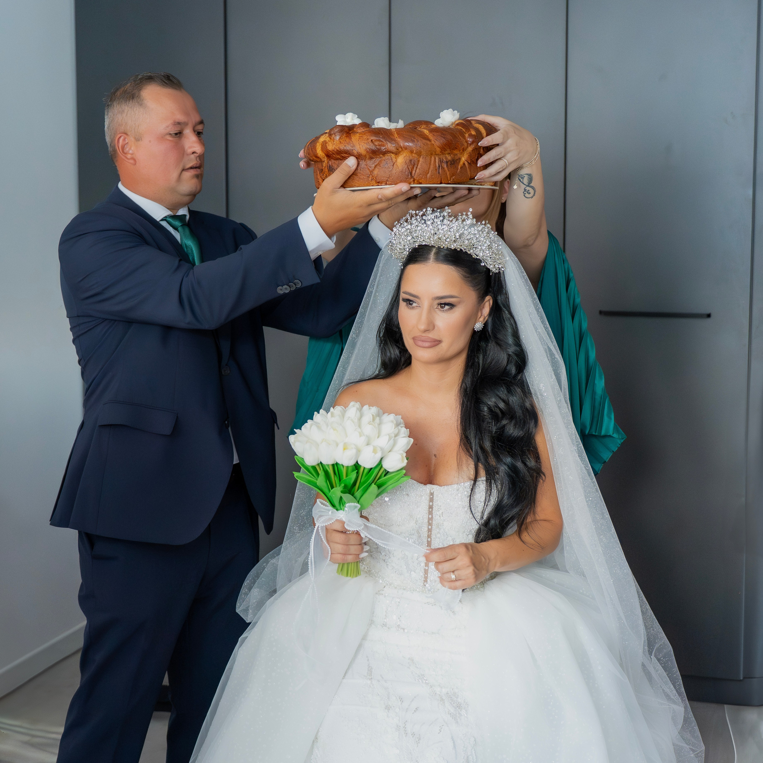 Bride in wedding gown holding white bouquet with bread held above her head by two people.