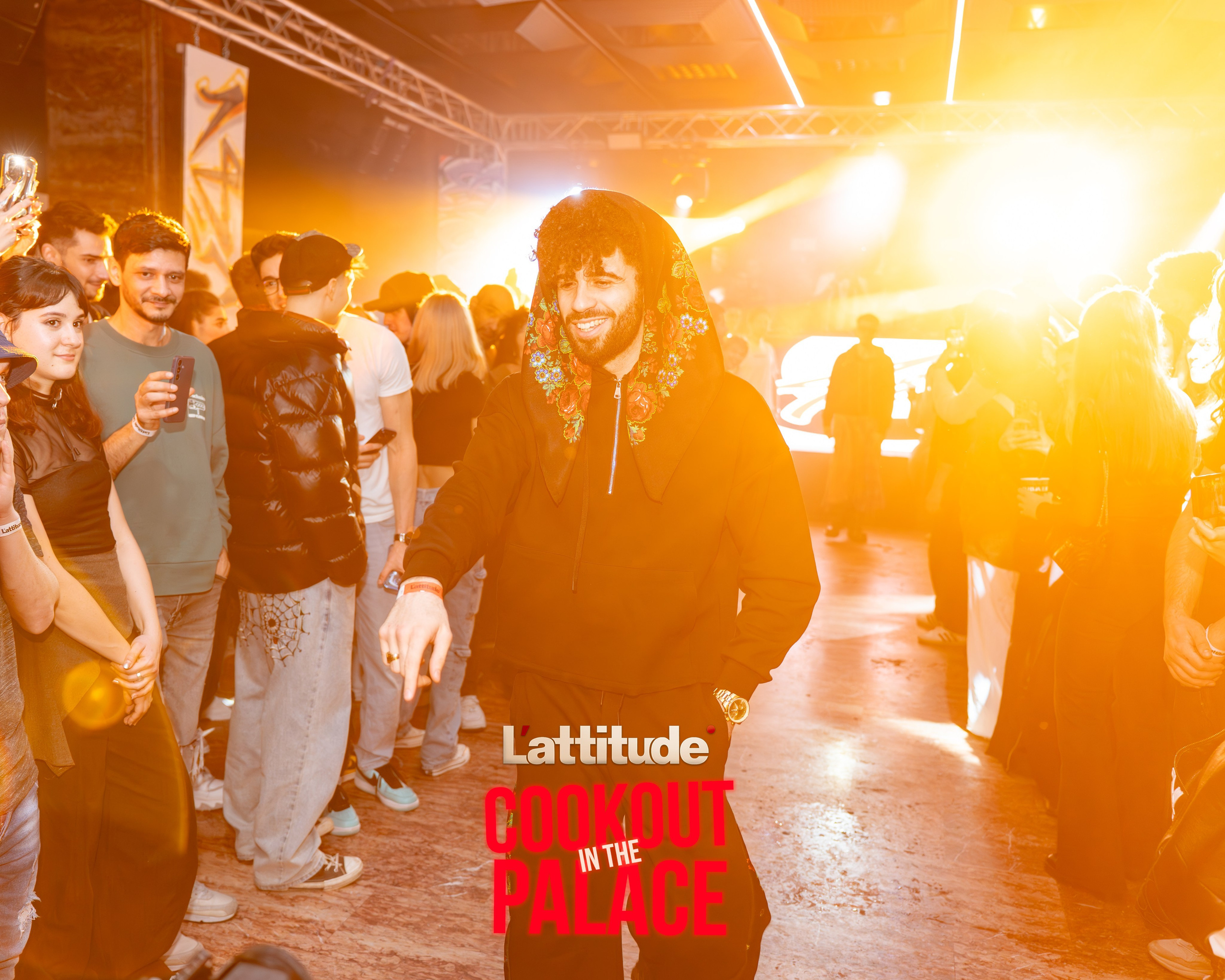 Man in black hoodie with floral hood smiling and pointing at an indoor event with crowd and bright stage lights.