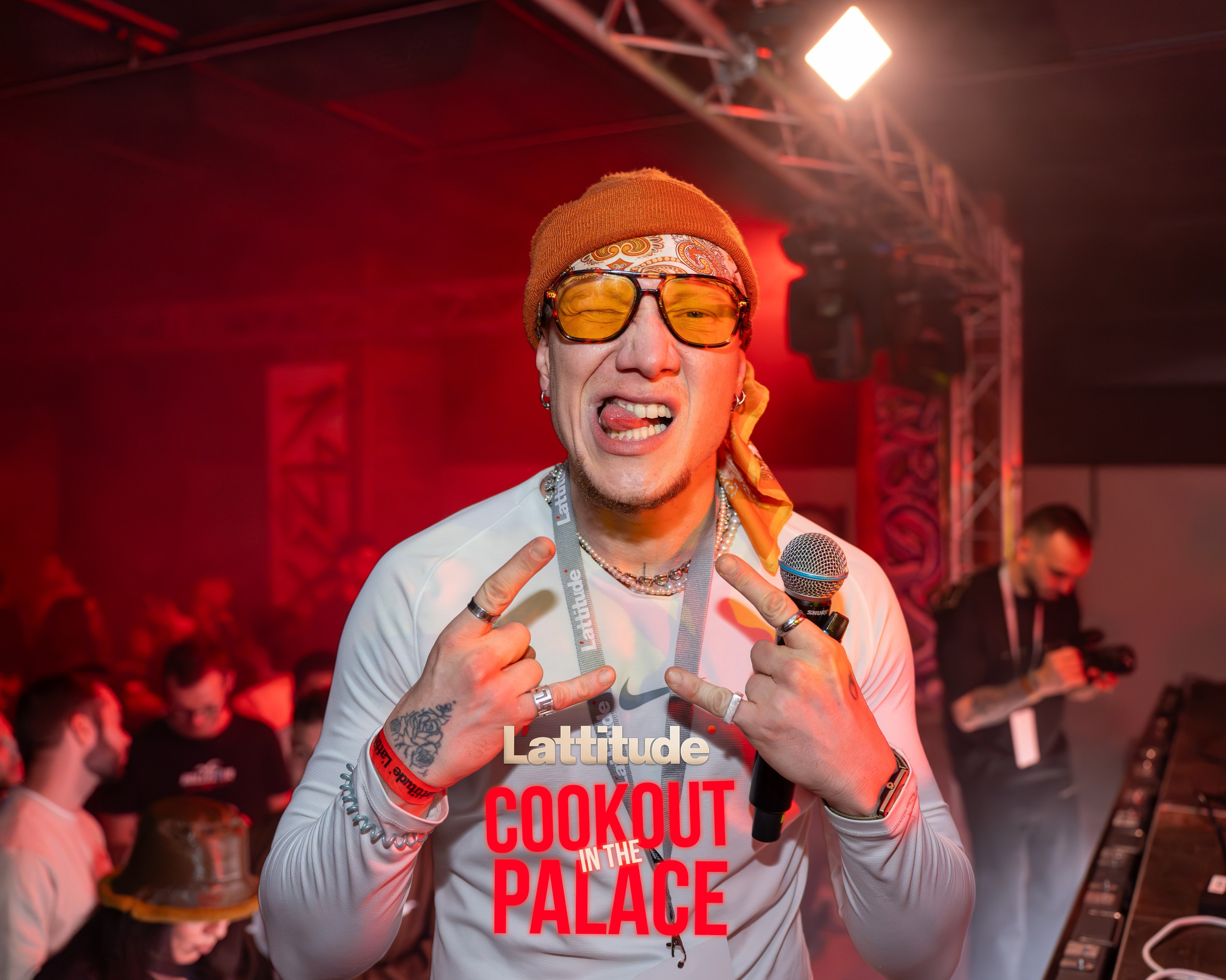 Man in orange beanie and yellow glasses making hand signs and holding microphone at event.