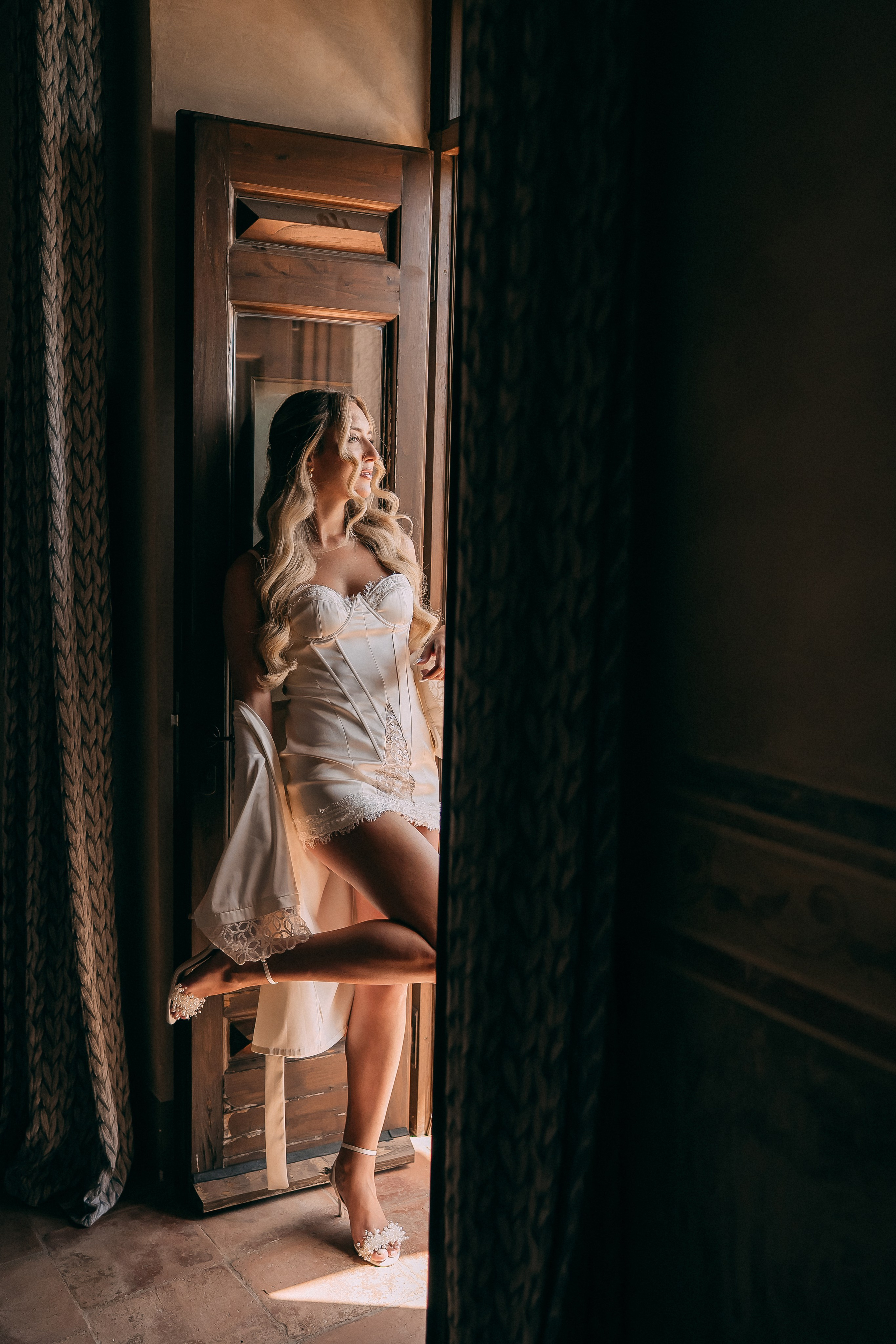 Bride in a silk robe standing by the window in soft morning light