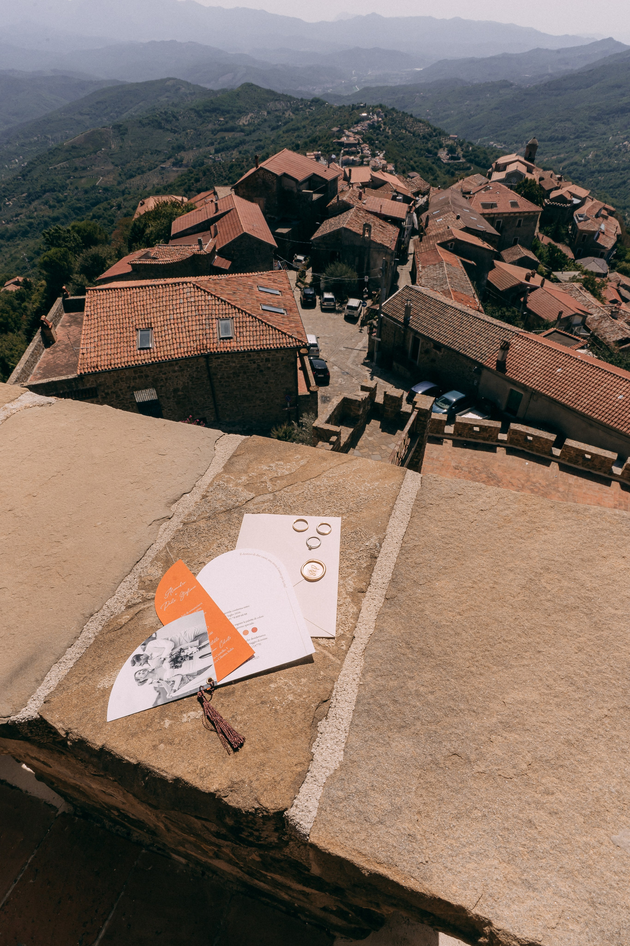 Wedding invitation suite and rings on a stone ledge above the village rooftops