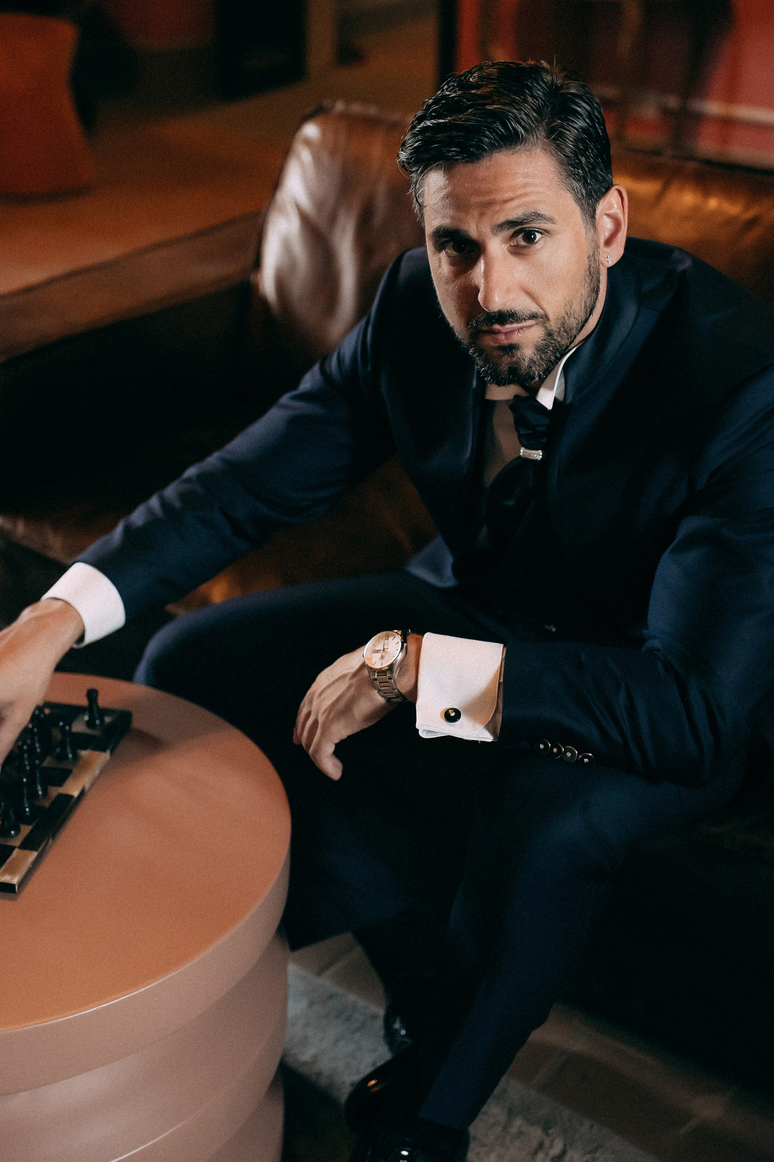Groom in a navy tuxedo sitting by a chess table before the ceremony