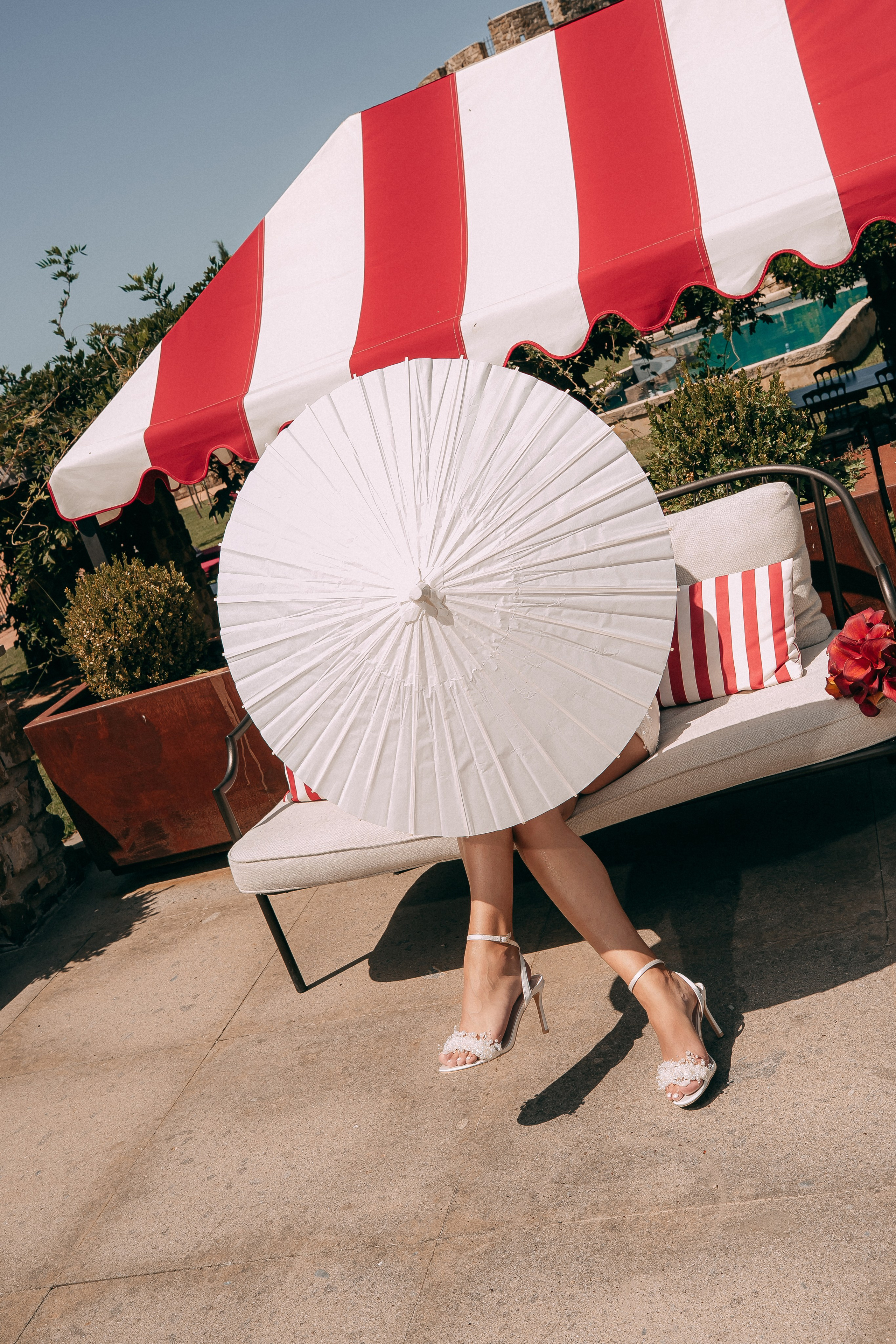 Bride with a paper parasol under a red striped umbrella by the pool