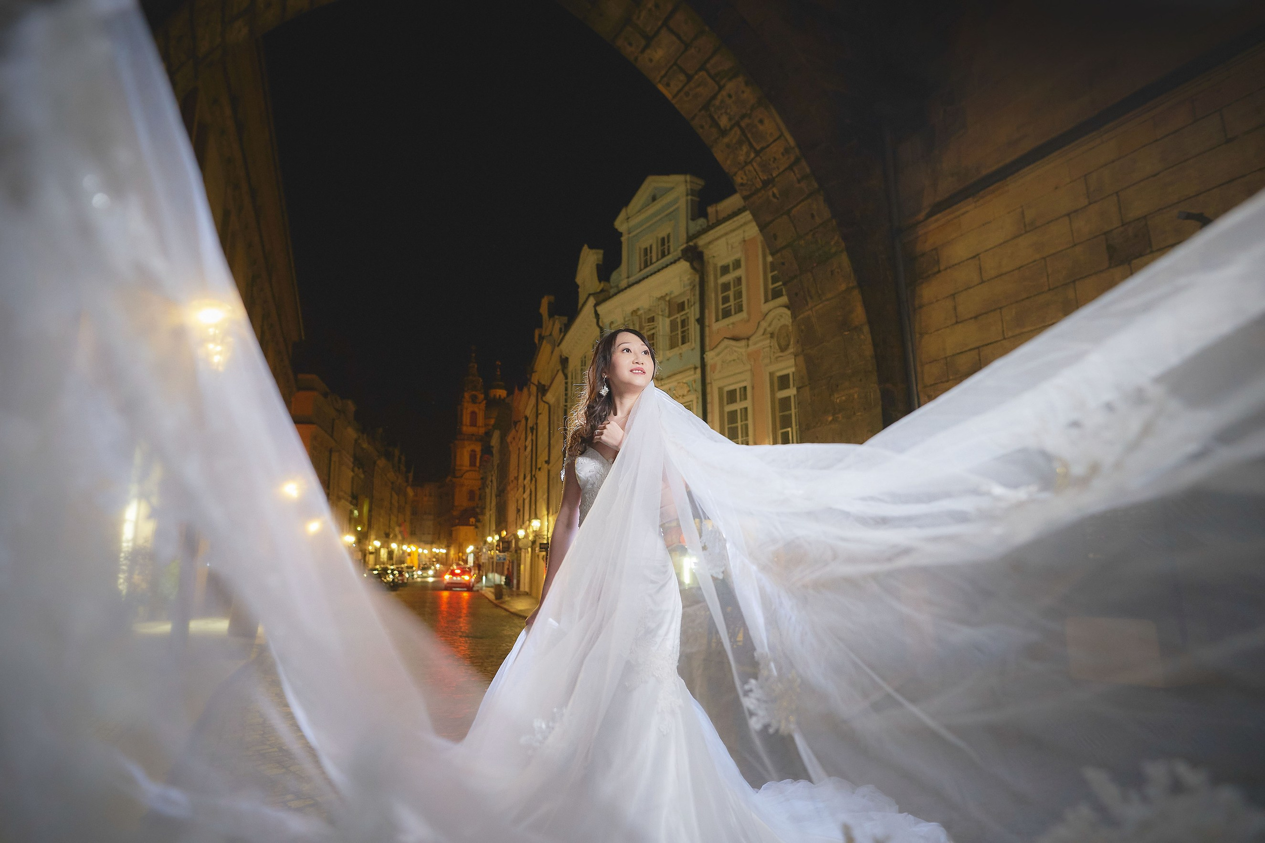 Bride Eva posing with two veils beneath a gothic tower archway in Prague at dawn.