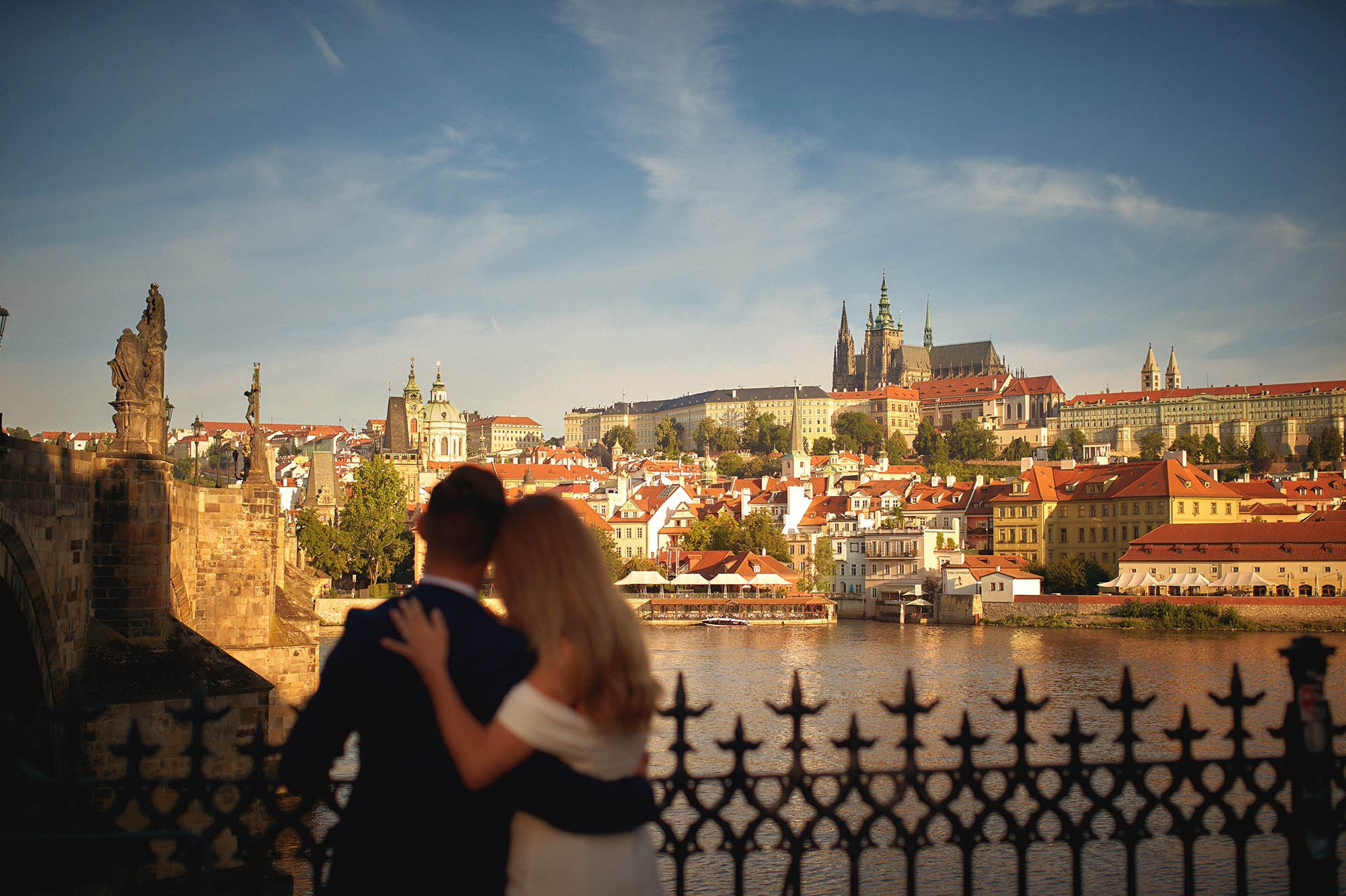 Couple embracing in near-silhouette as sunrise illuminates historic Prague Castle skyline.