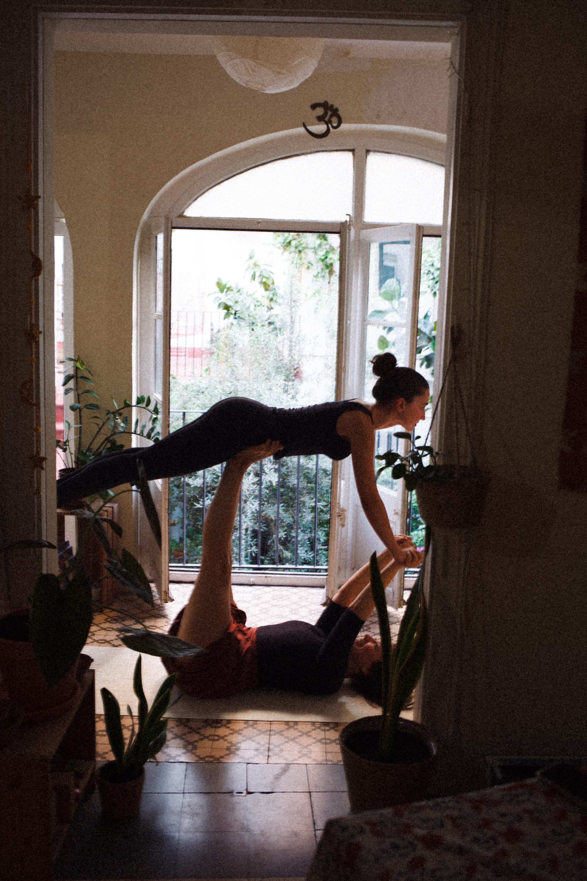Two women practice yoga inside a room lit by soft daylight from an open door to the garden