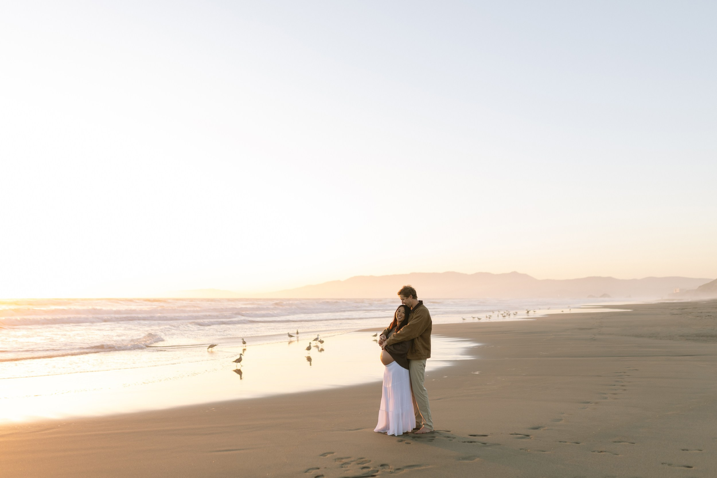 Pregnant woman in flowing dress standing by the Pacific Ocean at sunset