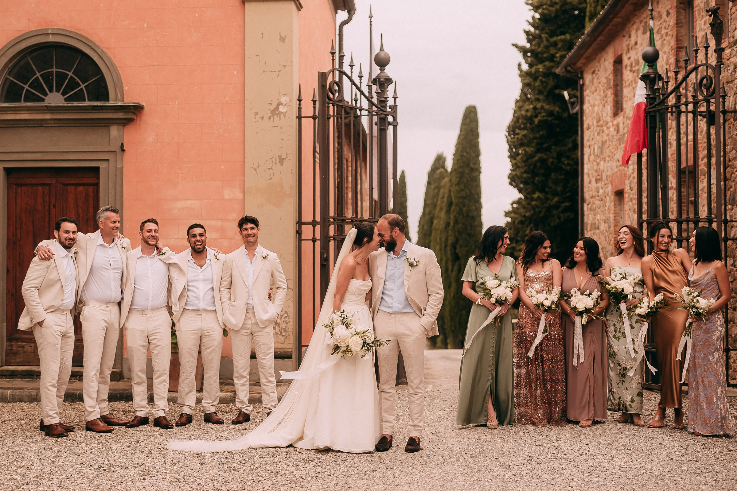 Bridal party and groomsmen with newlyweds pose together in elegant attire at the wedding venue entrance in Tuscany.