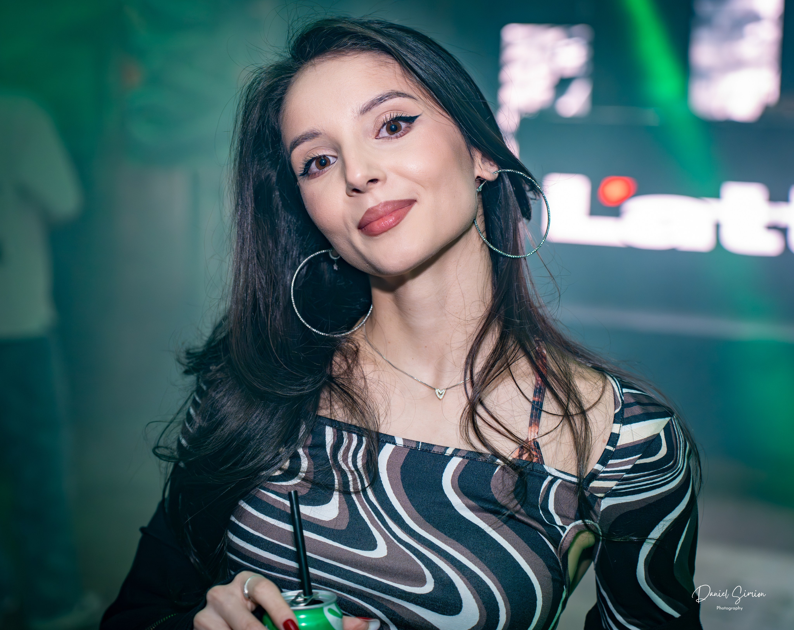 Young woman with hoop earrings and patterned top holding a drink, facing the camera indoors.