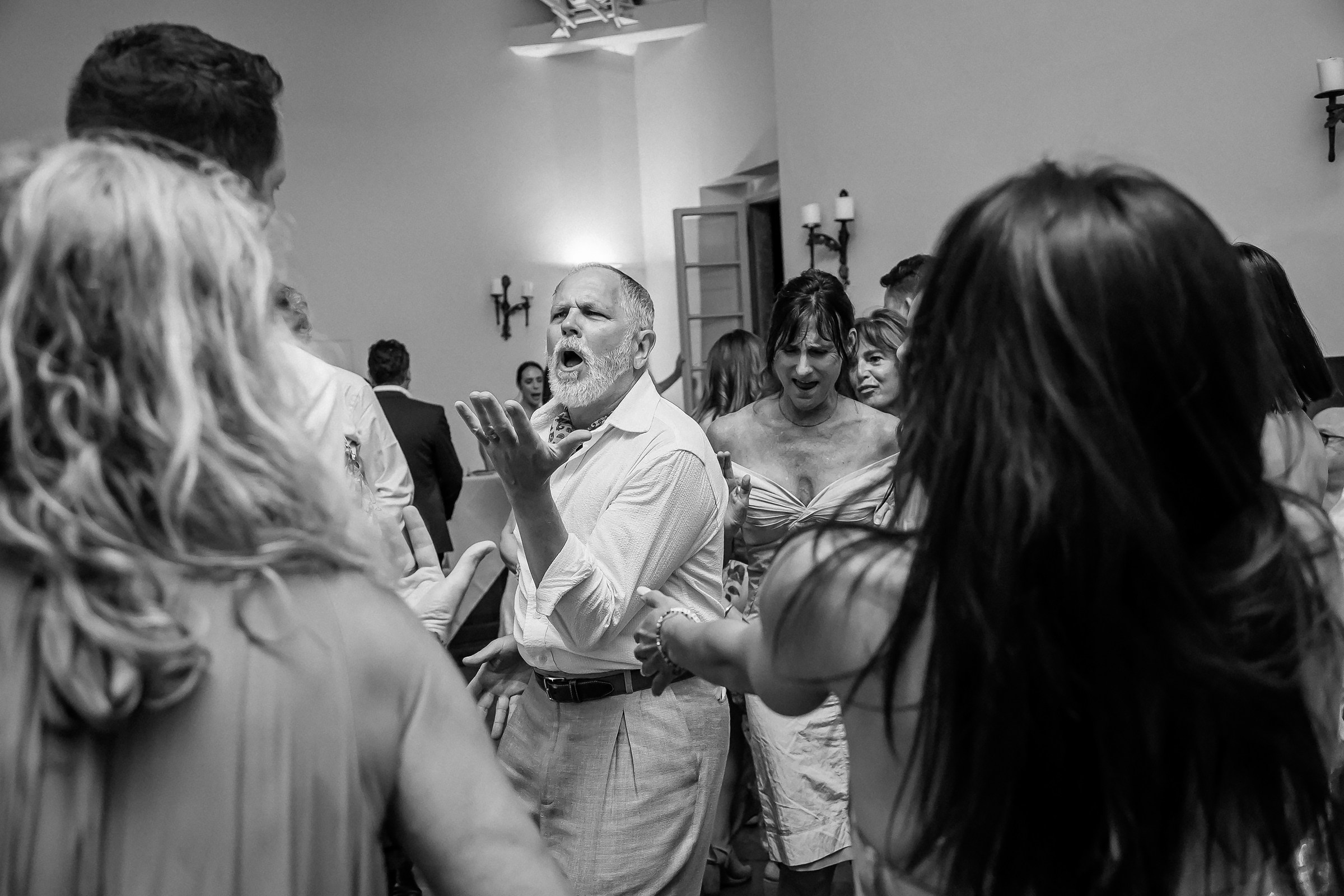 A man passionately singing and gesturing while surrounded by a lively crowd on a dance floor, capturing the energy of the celebration.