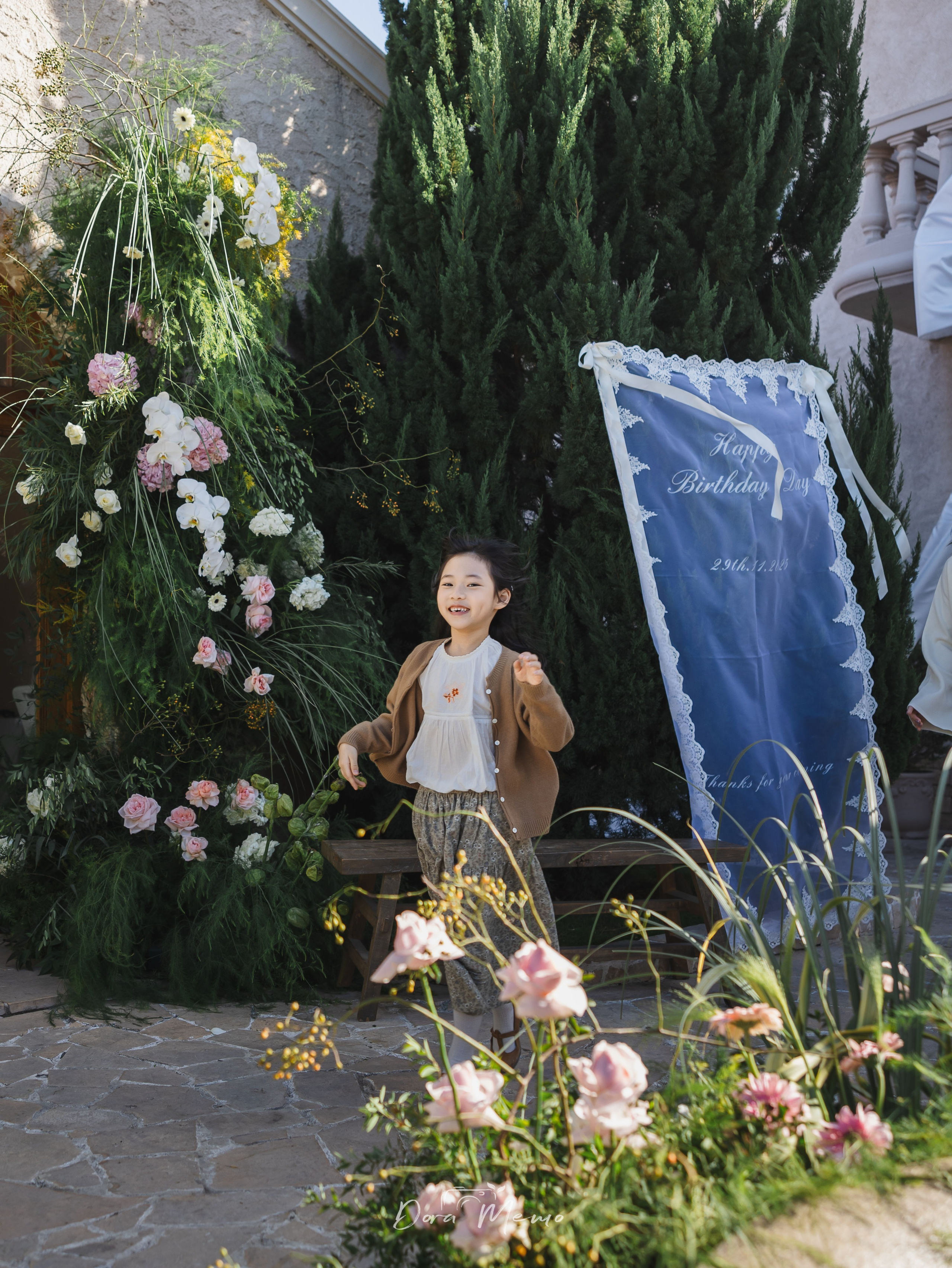 Young girl standing in front of flowers with gentle smile, outdoor birthday photography, documentary style