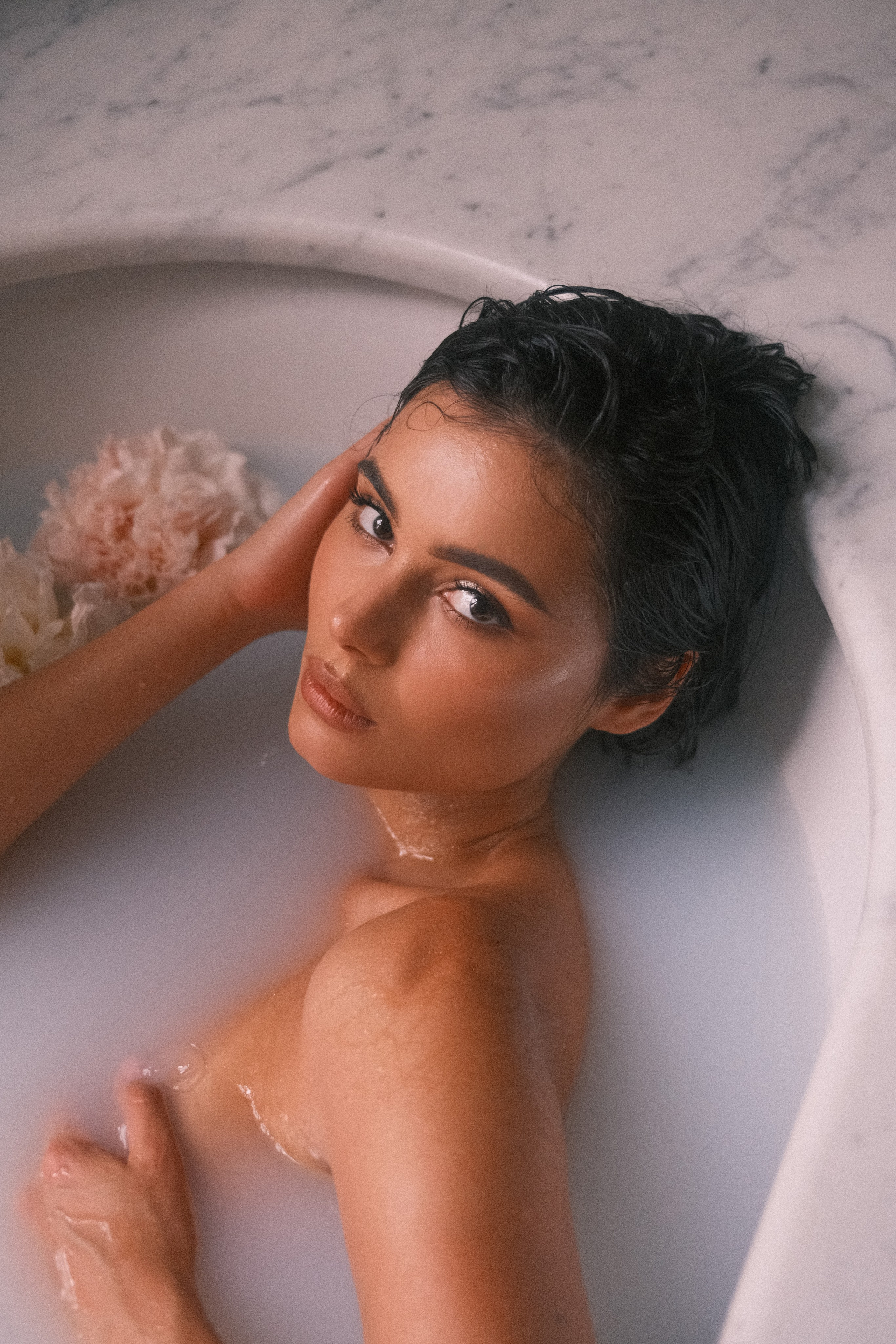 Close-up of a bride reclining in a bathtub, her confident gaze meeting the viewer’s, with her elegant features complemented by the soft ambiance of the flowers.