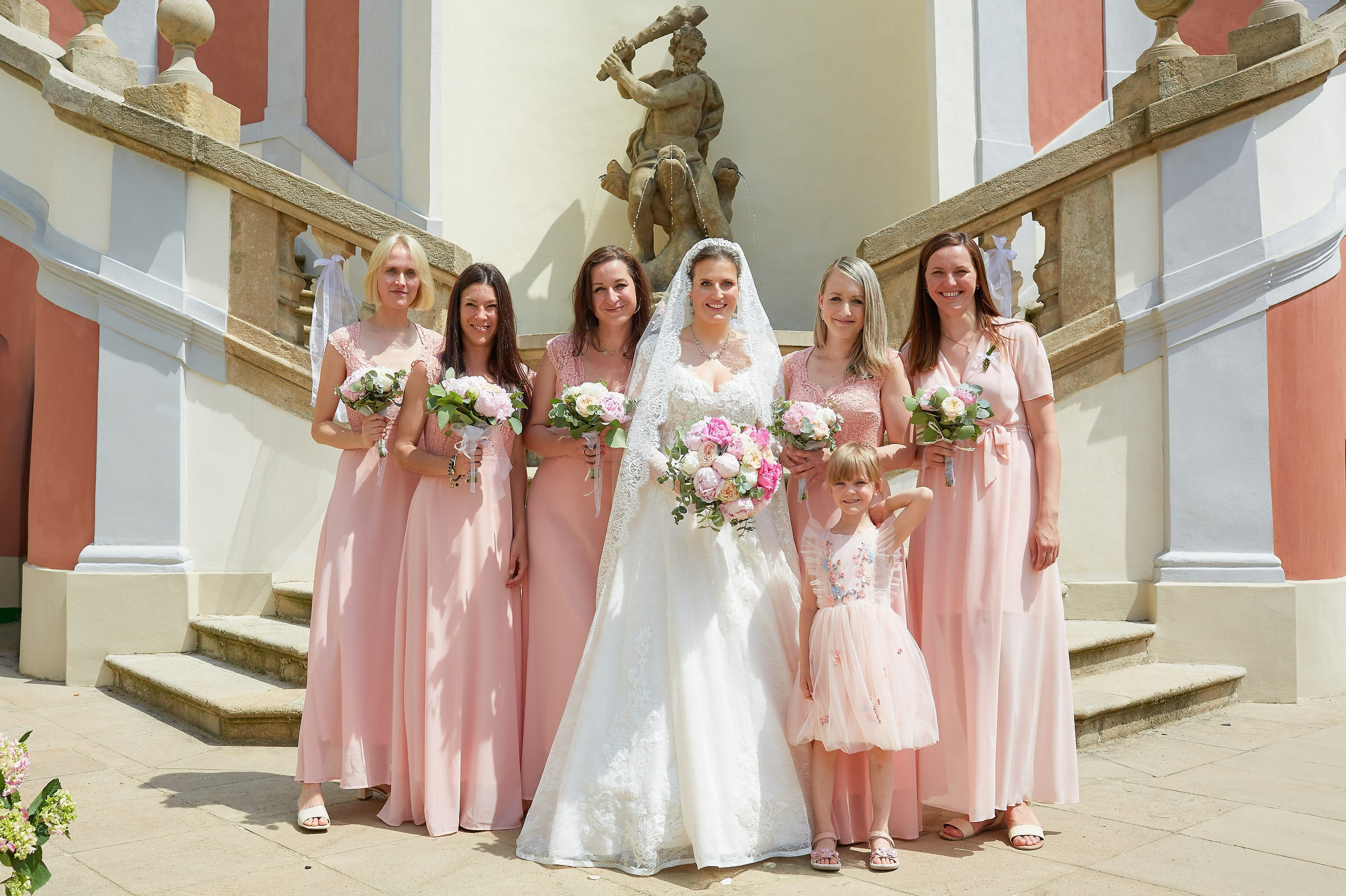 With a famous statue standing behind them, a beaming bride is surrounded by her rose-colored attired bridesmaids and a young flower girl on the steps of the historic Ledebour Garden.