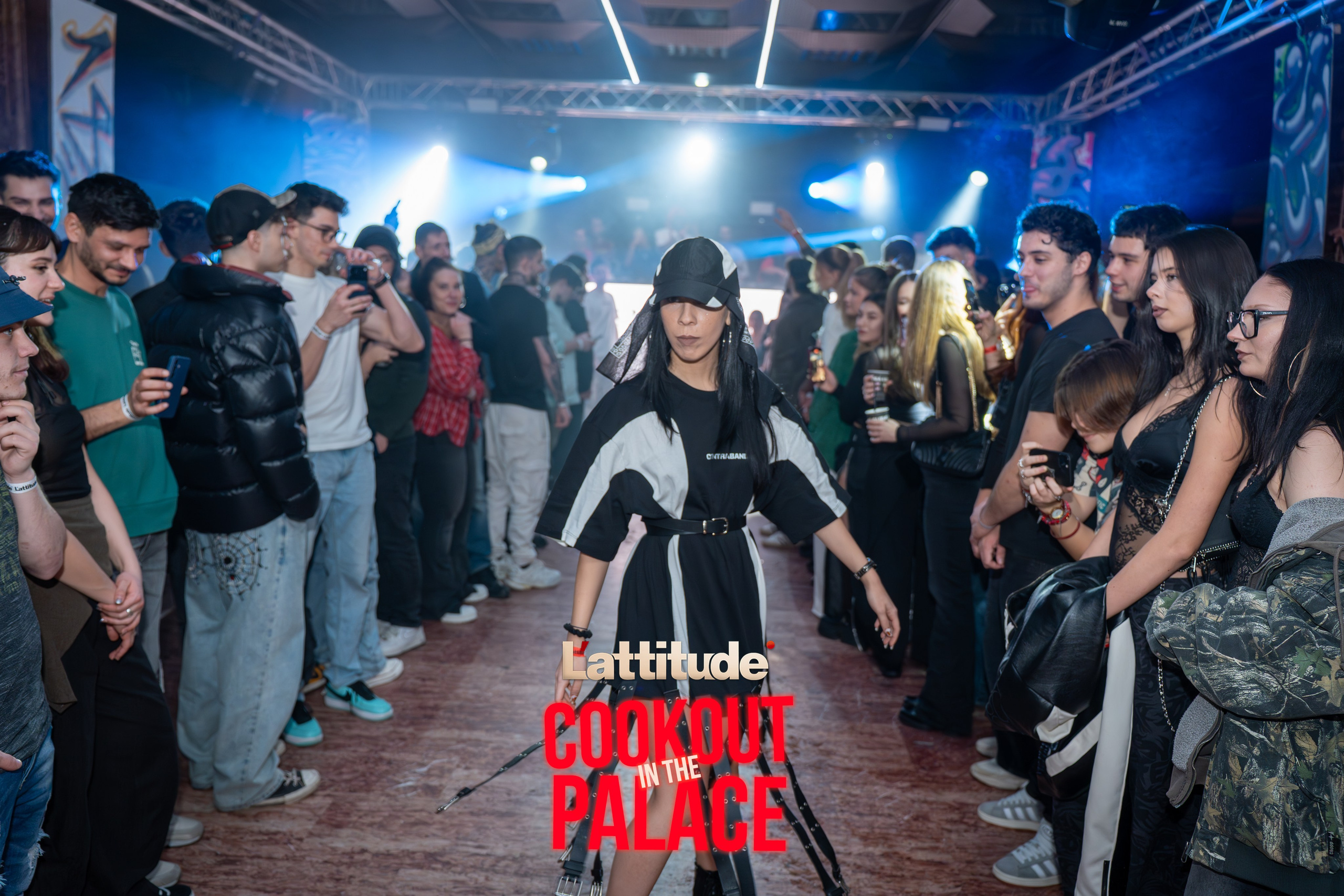Woman in black and white outfit walking between crowd watching at indoor event with stage lighting.