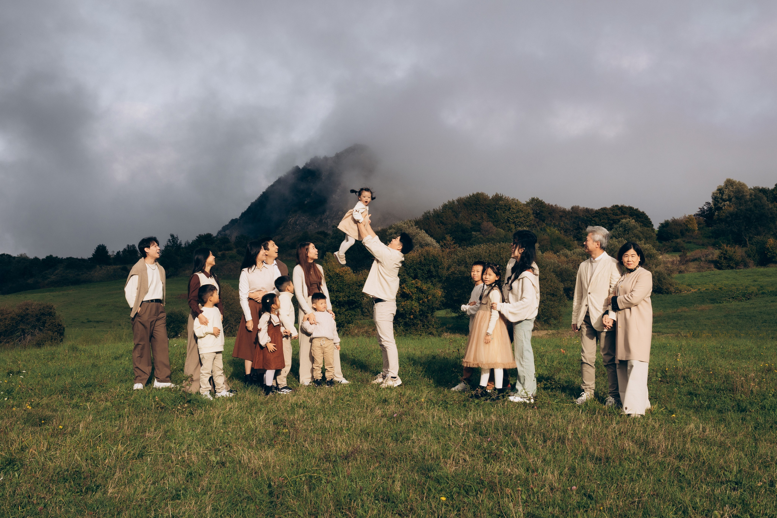 Grande famiglia con bambini su un prato verde tra le montagne di San Marino, più generazioni insieme, padre che solleva un bambino in aria, luce naturale, cielo nuvoloso, fotografia di famiglia spontanea all’aperto.