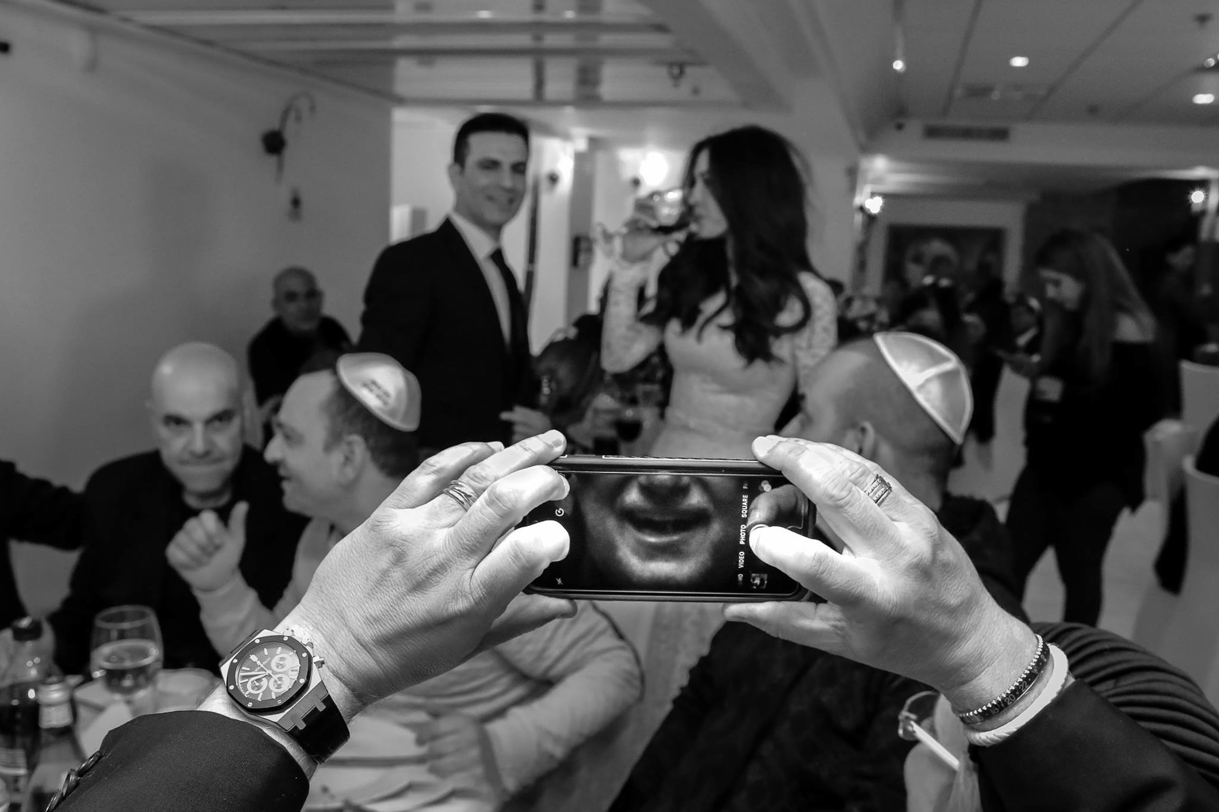 A wedding guest holds a smartphone in front of his face while the bride and groom stand in the background