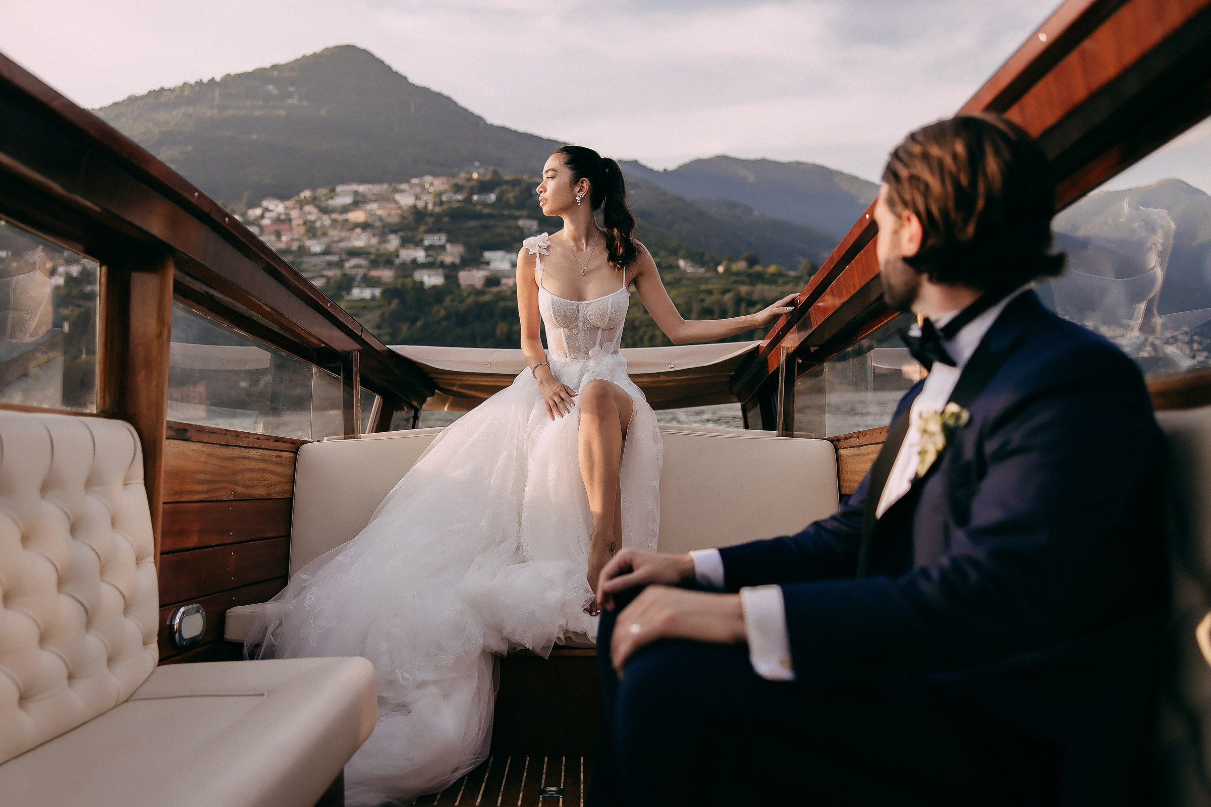 Bride poses gracefully on boat, groom watches her against mountain view.
