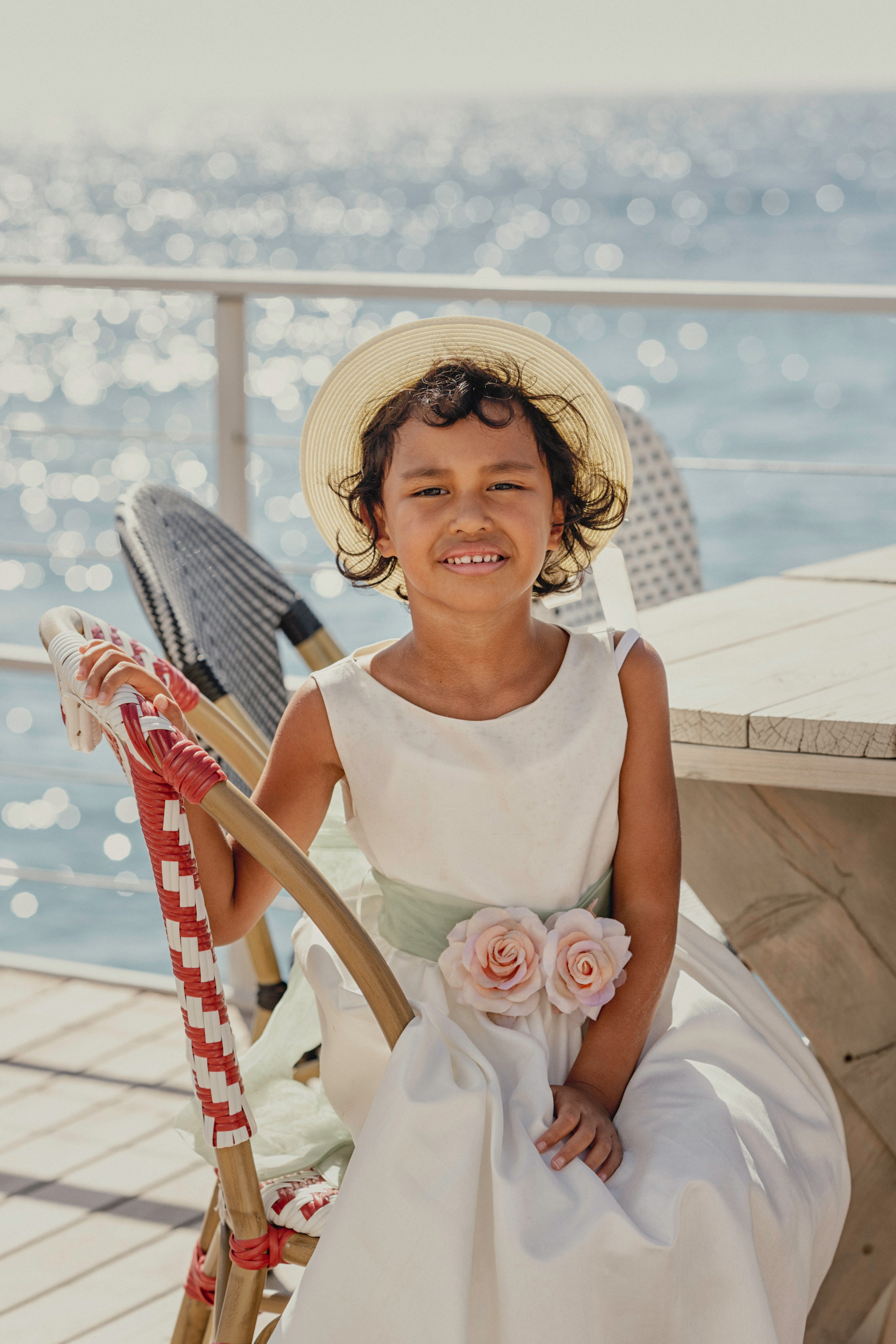 Une fille en robe et chapeau assise sur une chaise, avec vue sur la mer.