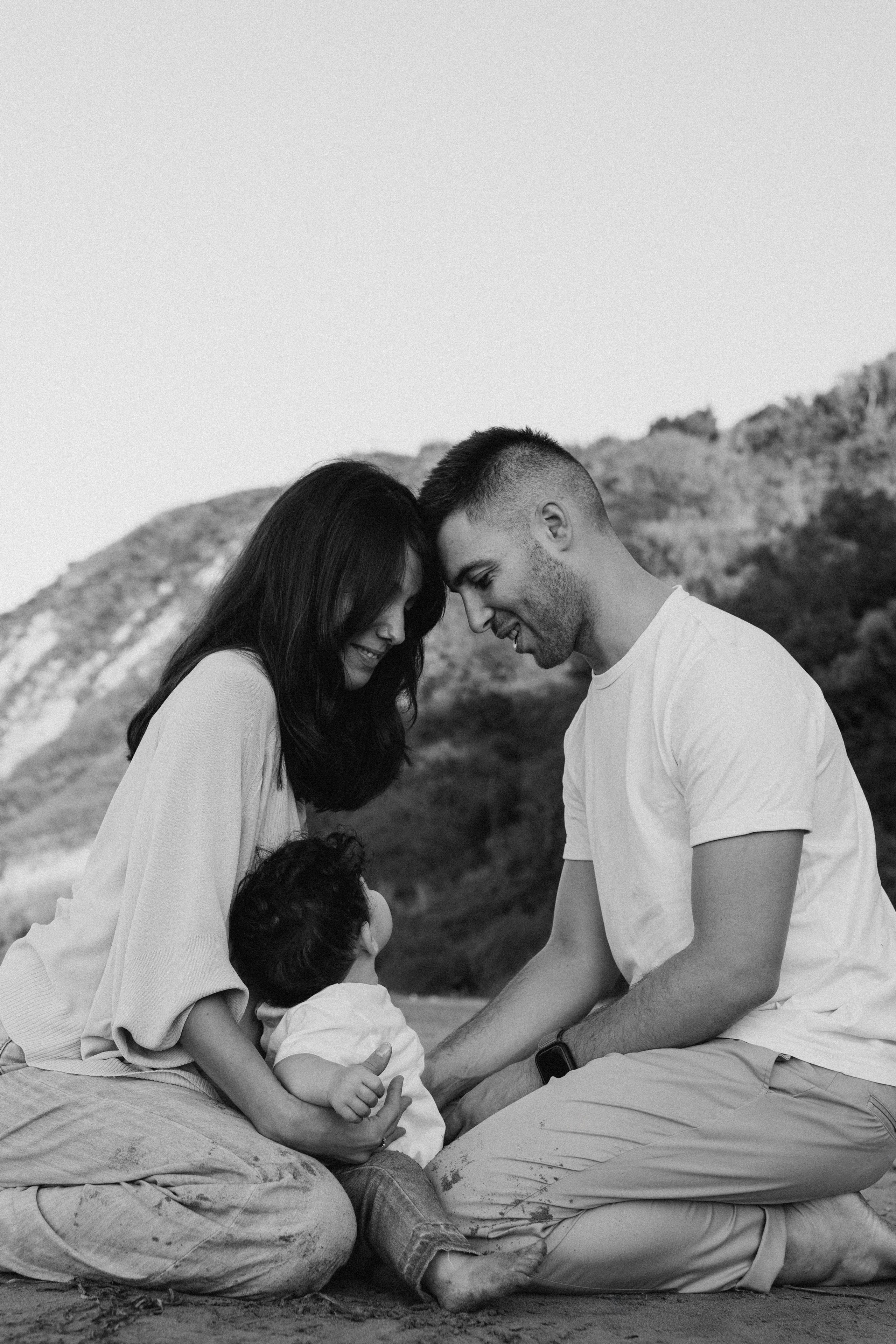 Ritratto di famiglia in bianco e nero sulla spiaggia di Cattolica: i genitori sono seduti sulla sabbia di fronte al loro bambino, piegati verso di lui, in un’atmosfera intima e silenziosa, con il paesaggio naturale sullo sfondo che rafforza il senso di vicinanza.